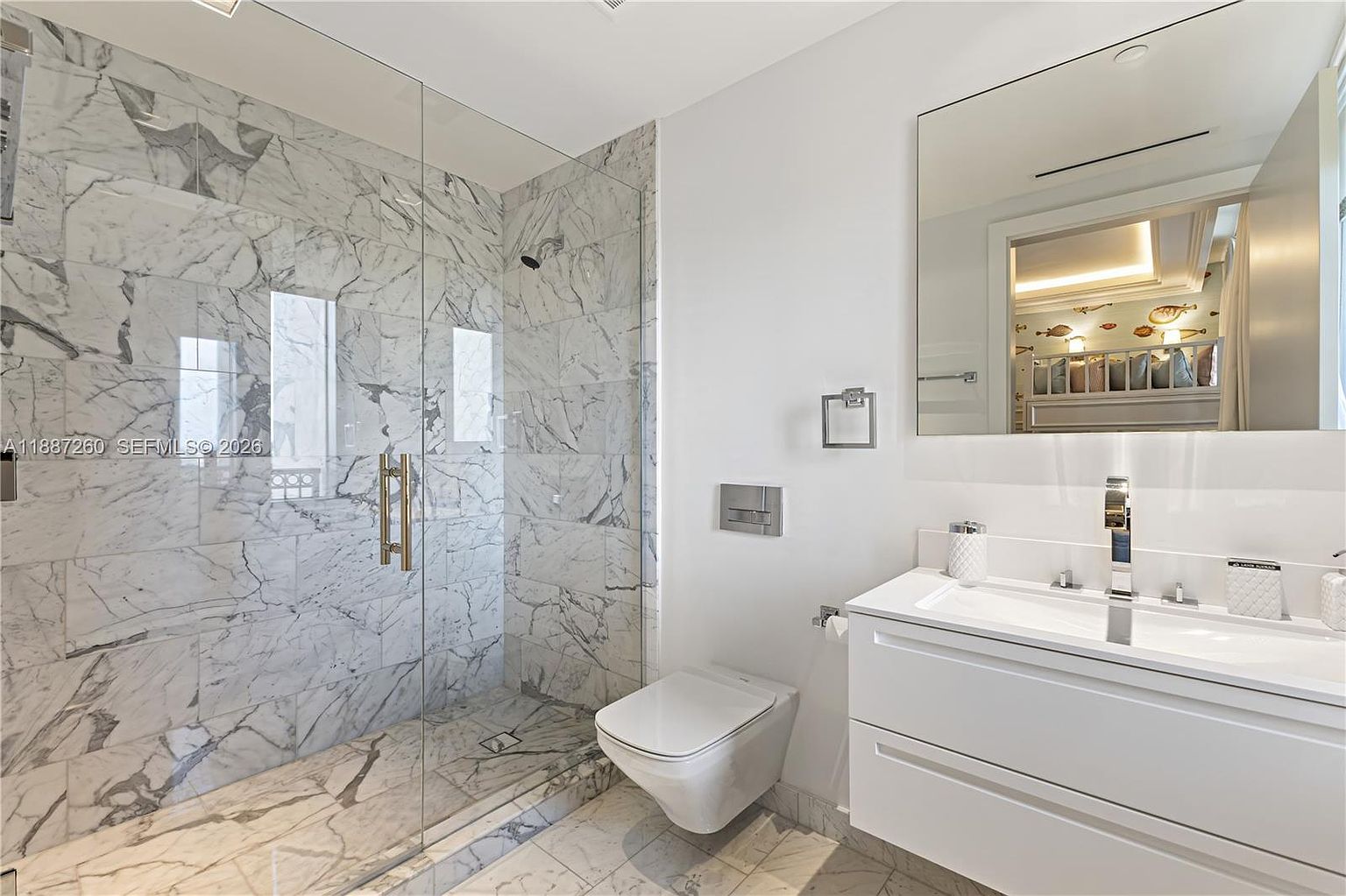 This modern, high-end bathroom features striking floor-to-ceiling marble tile with dramatic veining throughout the shower and flooring. The space is equipped with a sleek wall-mounted toilet, a minimalist white vanity with a rectangular sink, and a large frameless glass shower enclosure. The bright, clean aesthetic is enhanced by a large mirror reflecting a glimpse of an adjacent room, creating a sophisticated and luxurious atmosphere.
