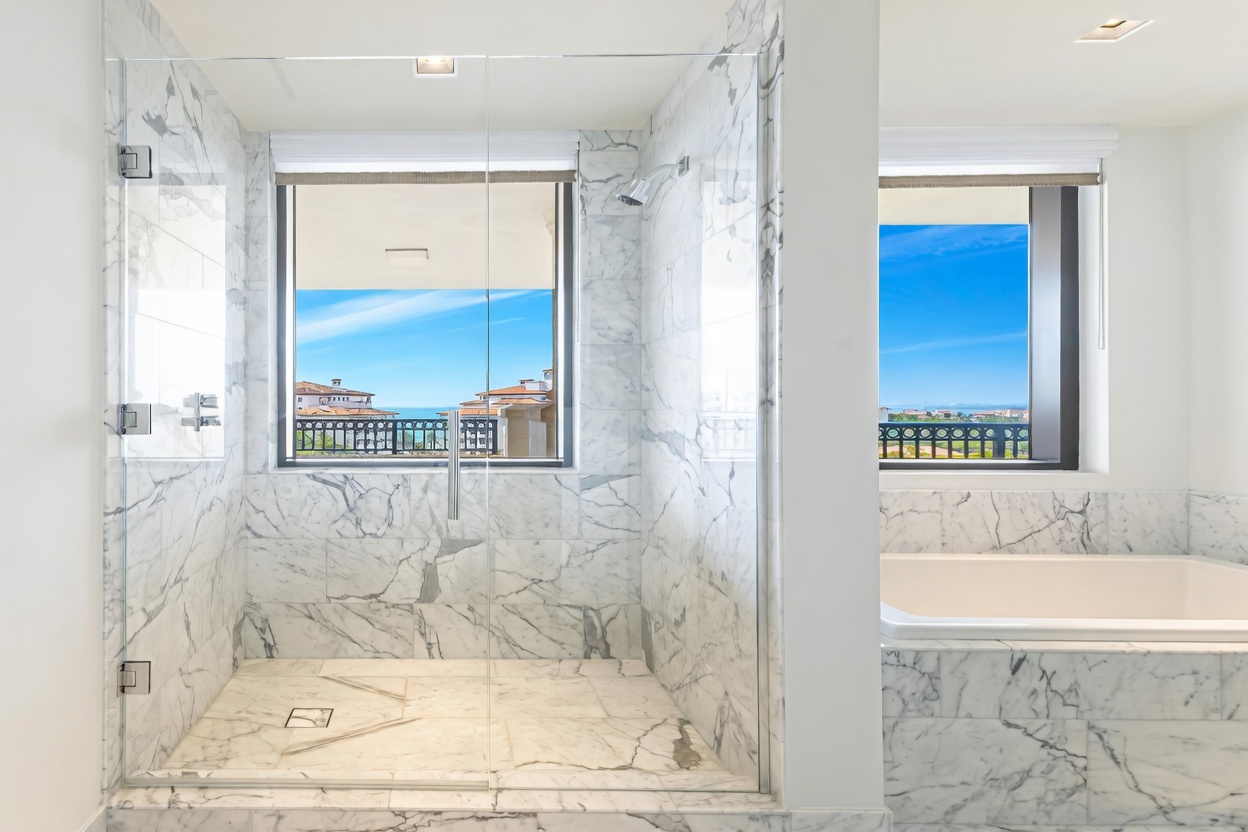 This luxurious primary bathroom features a spacious walk-in glass shower and a deep soaking tub, both adorned with elegant white marble tile with grey veining. Large windows above both the shower and tub provide abundant natural light and stunning, unobstructed views of the ocean and surrounding landscape. The overall aesthetic is sophisticated, bright, and spa-like, emphasizing high-end finishes and a seamless connection to the outdoors.