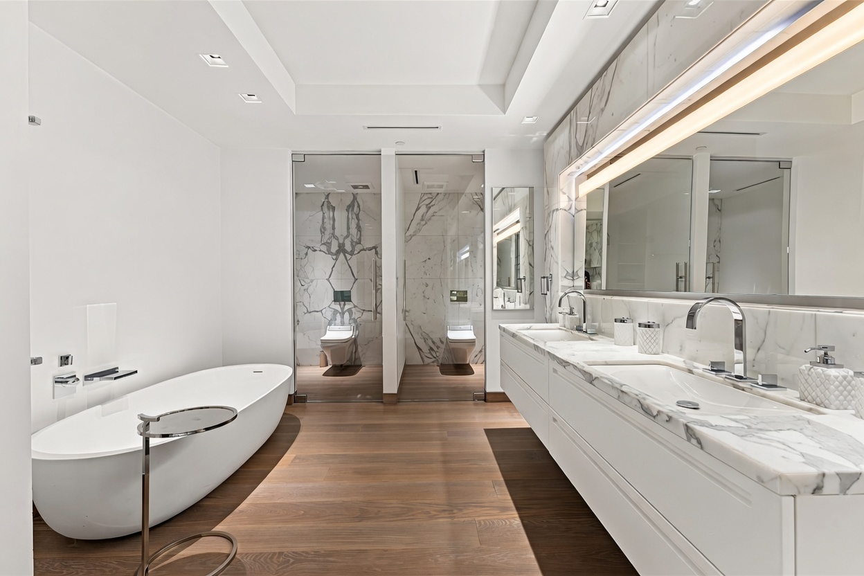 This luxurious primary bathroom features a sleek, modern design with a freestanding soaking tub and a long, double-sink vanity topped with white marble. The space is accented by warm wood flooring, floor-to-ceiling marble wall cladding, and two private, glass-enclosed toilet rooms. The bright, minimalist aesthetic is enhanced by recessed lighting and a large, integrated mirror, creating an atmosphere of high-end sophistication.