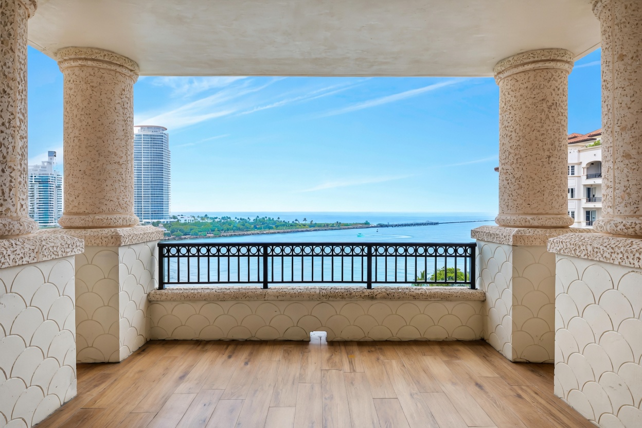 This elegant, covered balcony offers a stunning, unobstructed panoramic view of the ocean and distant city skyline. The space is framed by classic, textured stone columns and features a decorative, scalloped-patterned wall base and a sleek black metal railing. The perspective is centered, drawing the viewer's eye directly toward the serene horizon, creating a luxurious and tranquil atmosphere.