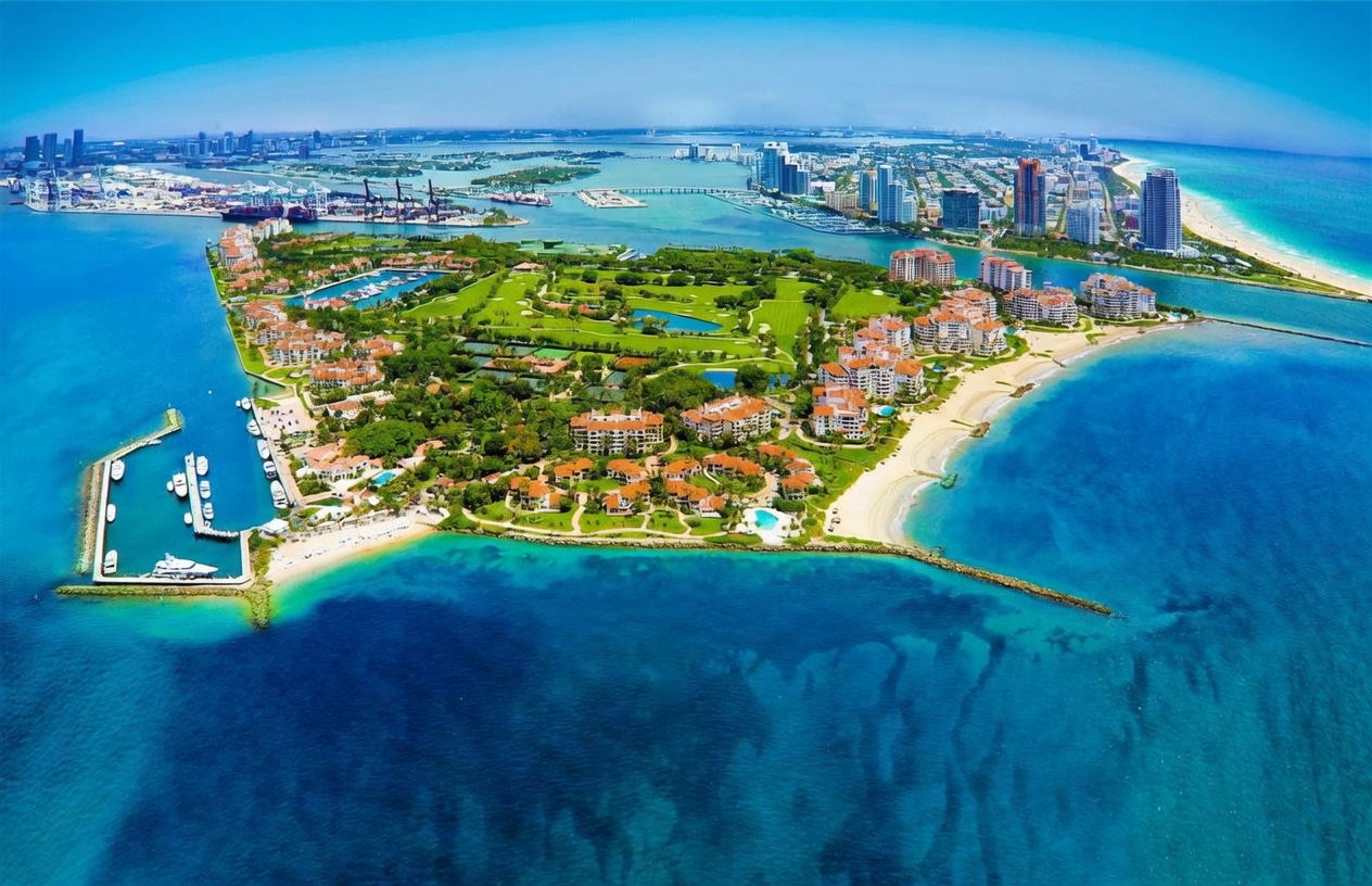 This stunning aerial perspective captures Fisher Island, a luxurious private enclave in Miami, showcasing its lush golf course, Mediterranean-style residential buildings, and pristine sandy beaches. The image highlights the island's exclusive waterfront location, surrounded by turquoise waters and offering a panoramic view of the distant Miami skyline and port. The composition emphasizes the island's serene, resort-like atmosphere and its unique integration of nature and high-end architecture.