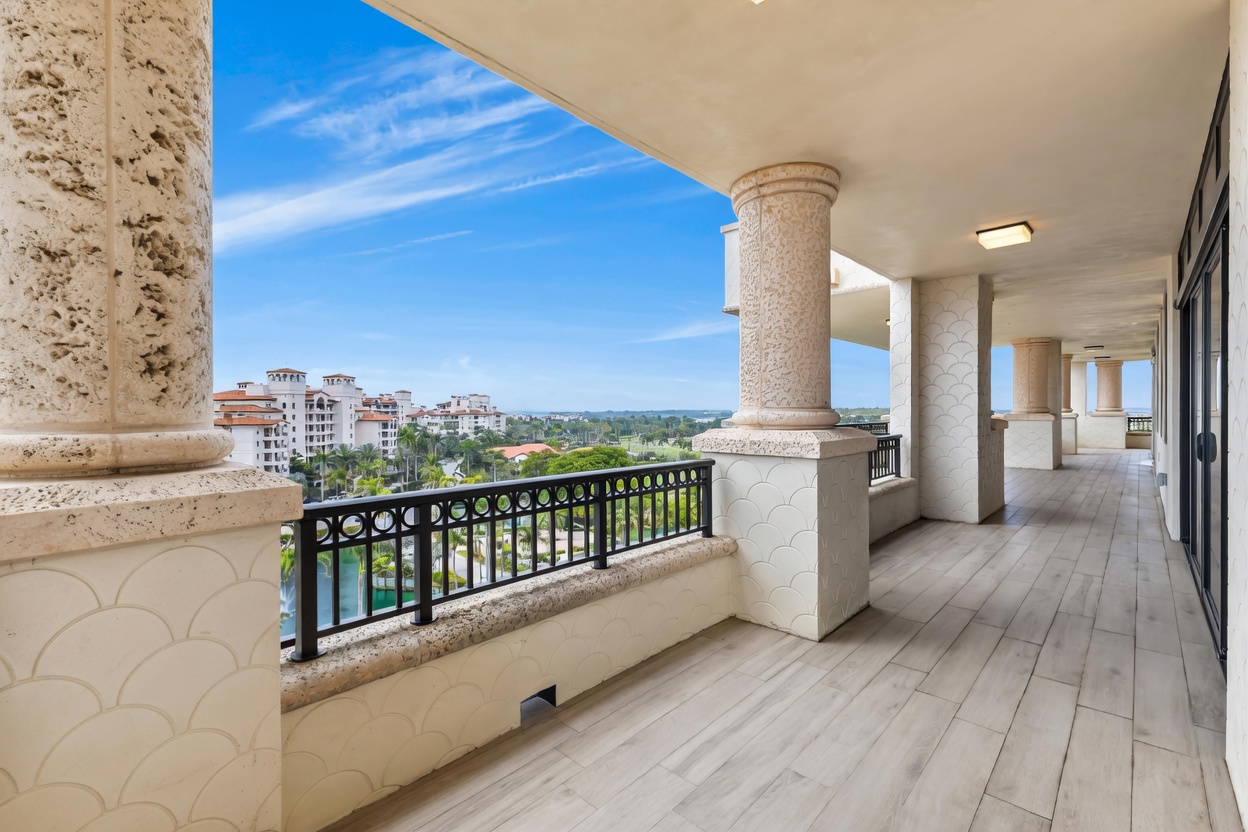 This expansive, covered balcony features light-toned wood-look tile flooring and elegant stone columns, creating a sophisticated outdoor living space. A black metal railing provides safety while maintaining an unobstructed, panoramic view of the surrounding resort-style community, lush greenery, and the distant ocean. The perspective is wide and inviting, emphasizing the seamless transition between the indoor living area and the scenic outdoor environment.