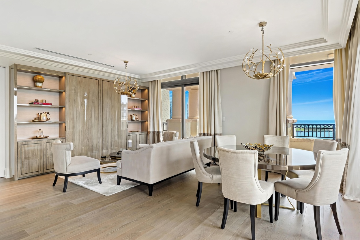 This elegant, open-concept living and dining area features a sophisticated neutral color palette with high-end finishes and light wood flooring. The space is anchored by a plush sofa and a circular dining table, complemented by unique, branch-like gold chandeliers and custom built-in cabinetry. Large floor-to-ceiling windows provide abundant natural light and a stunning view of the ocean, creating a luxurious and airy atmosphere.