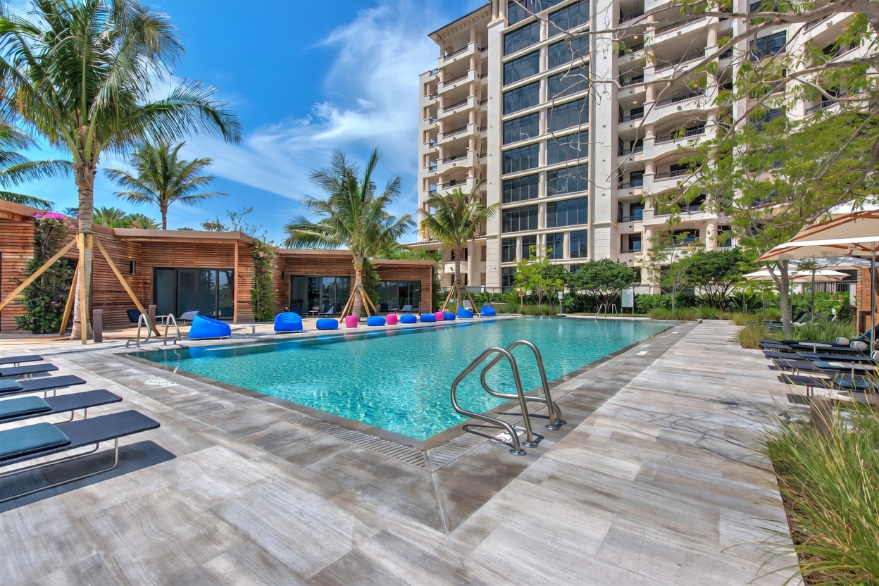 This vibrant outdoor pool area features a large, turquoise swimming pool surrounded by a light-toned stone deck and modern lounge chairs. The scene is framed by lush palm trees and a multi-story residential building in the background, creating a resort-like atmosphere. The perspective is a low-angle shot from the pool deck, emphasizing the inviting water and the clean, contemporary architectural design of the community amenities.