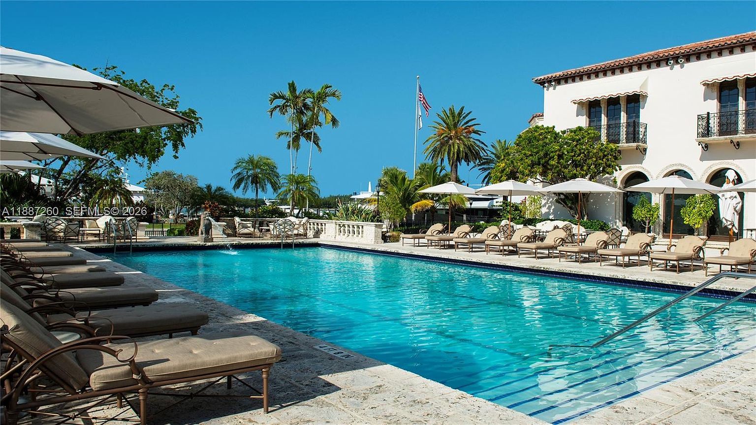 This image showcases a luxurious, expansive swimming pool set against a Mediterranean-style estate, featuring multiple lounge chairs and umbrellas arranged on a stone deck. The scene is framed by lush palm trees and a clear blue sky, creating a serene and high-end resort-like atmosphere. The perspective is from the poolside, looking toward the elegant white building, emphasizing the property's outdoor entertainment space.
