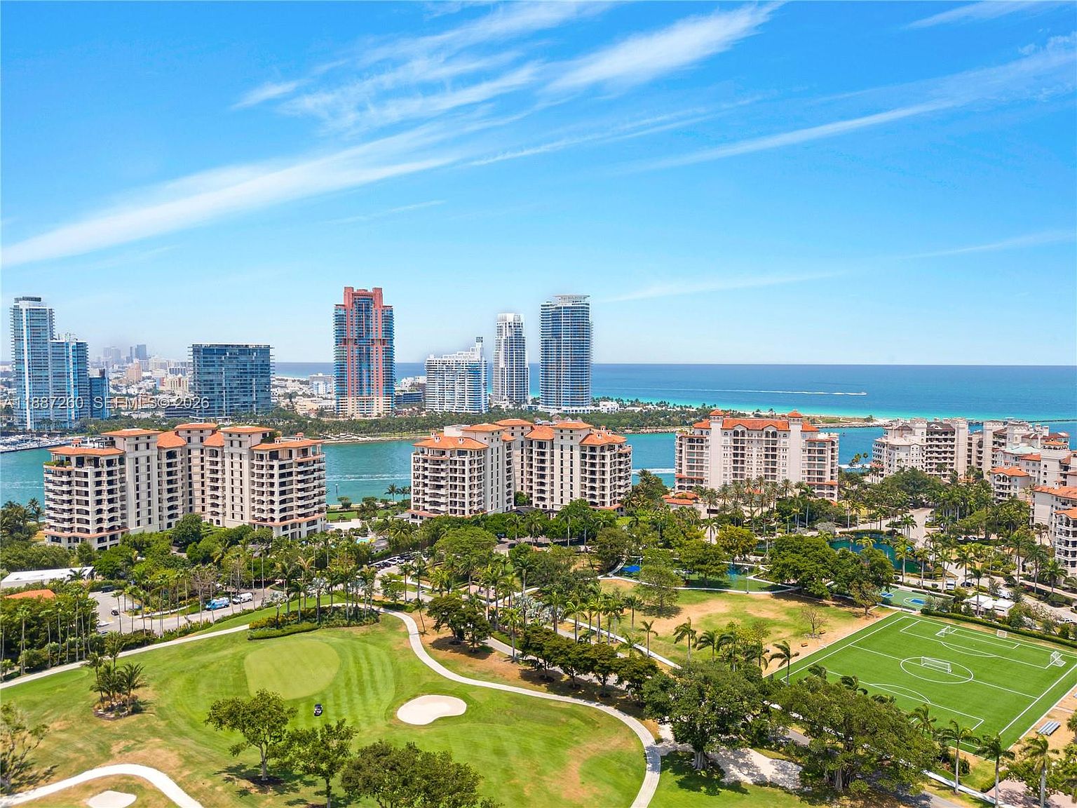 This high-angle aerial view captures a vibrant coastal landscape featuring a lush green golf course and a soccer field in the foreground. Beyond the park-like grounds, several mid-rise residential buildings overlook a serene waterway, with the expansive blue ocean and city skyline visible in the distance. The bright, sunny day highlights the scenic blend of urban development and recreational green space.