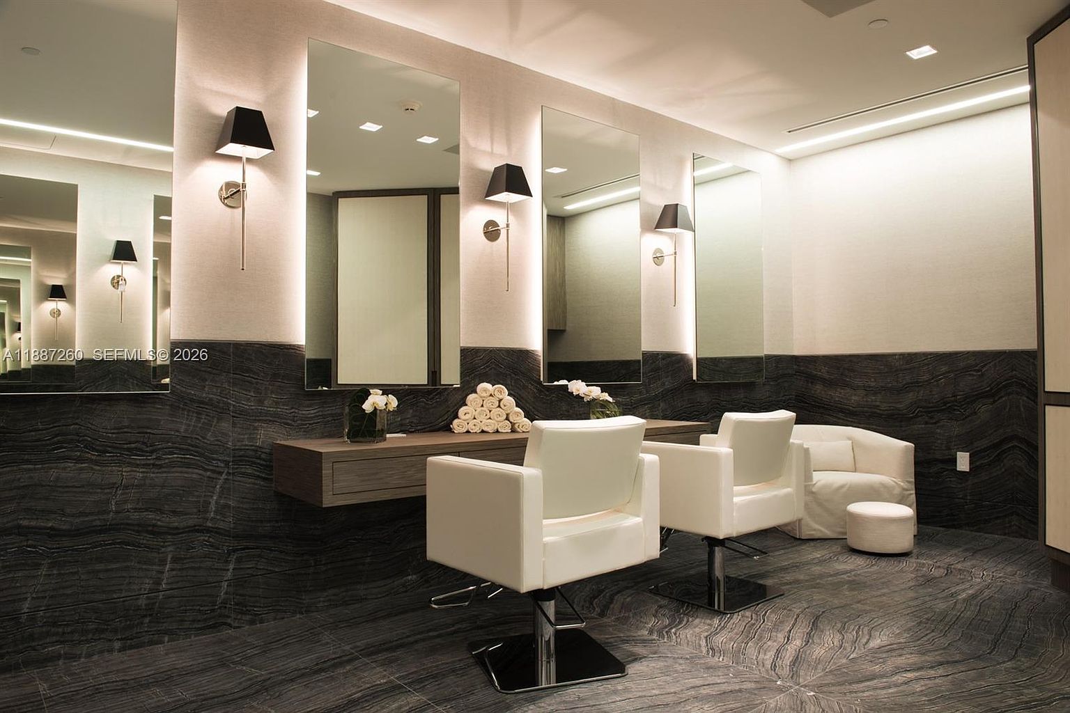 This sophisticated salon or spa amenity space features a sleek, modern design with dark, dramatic marble-patterned walls and flooring. Two white styling chairs are positioned in front of large, backlit mirrors, complemented by elegant wall sconces and a stack of neatly rolled towels on the vanity. The overall aesthetic is luxurious and serene, suggesting a high-end residential building or resort facility.