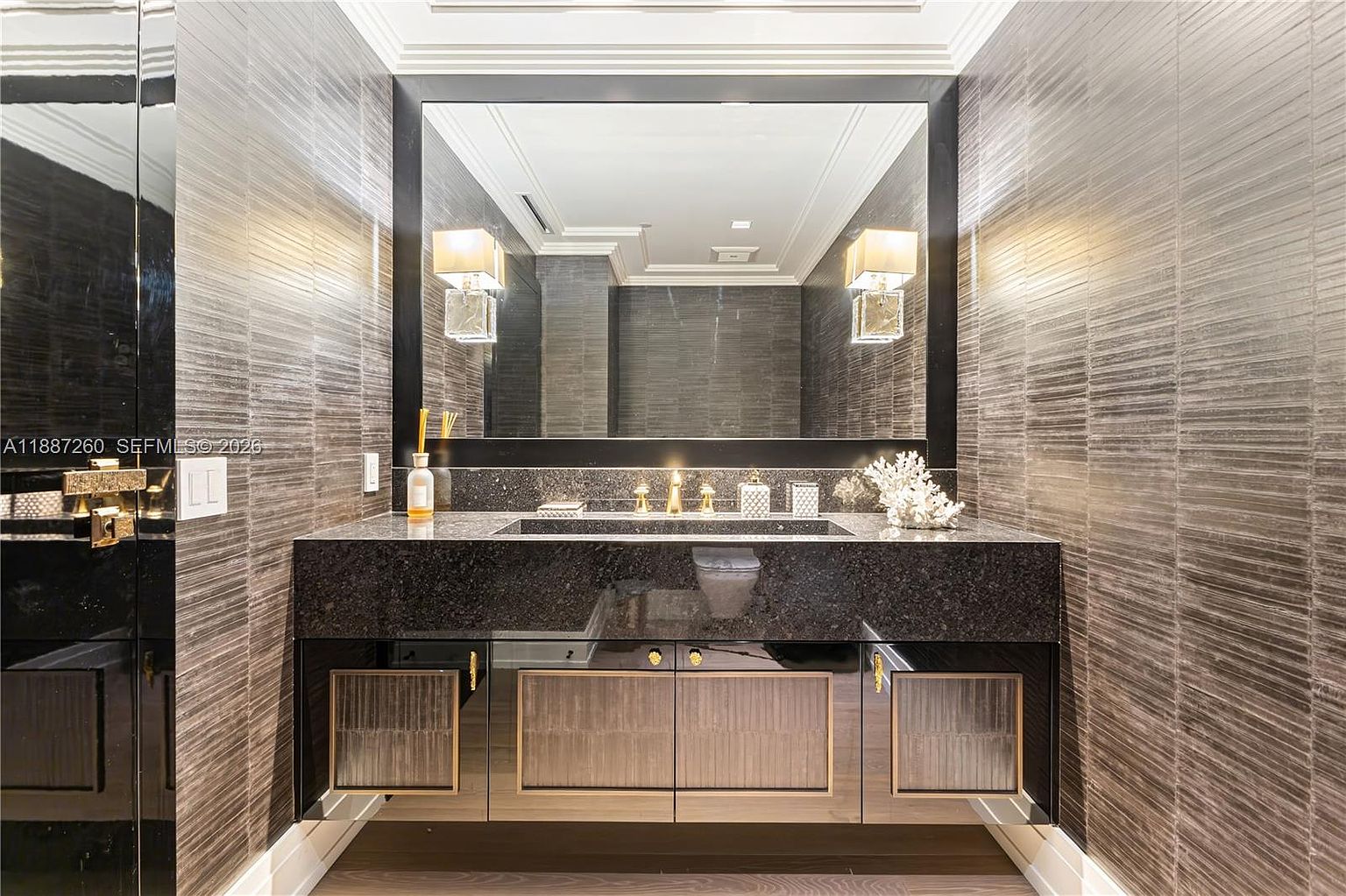 This sophisticated powder room features a striking dark granite vanity with a sleek integrated sink and gold-toned fixtures. The walls are adorned with textured, horizontal-patterned wallpaper, complemented by a large, dark-framed mirror and elegant wall sconces. The overall aesthetic is modern, luxurious, and moody, creating a high-end atmosphere.