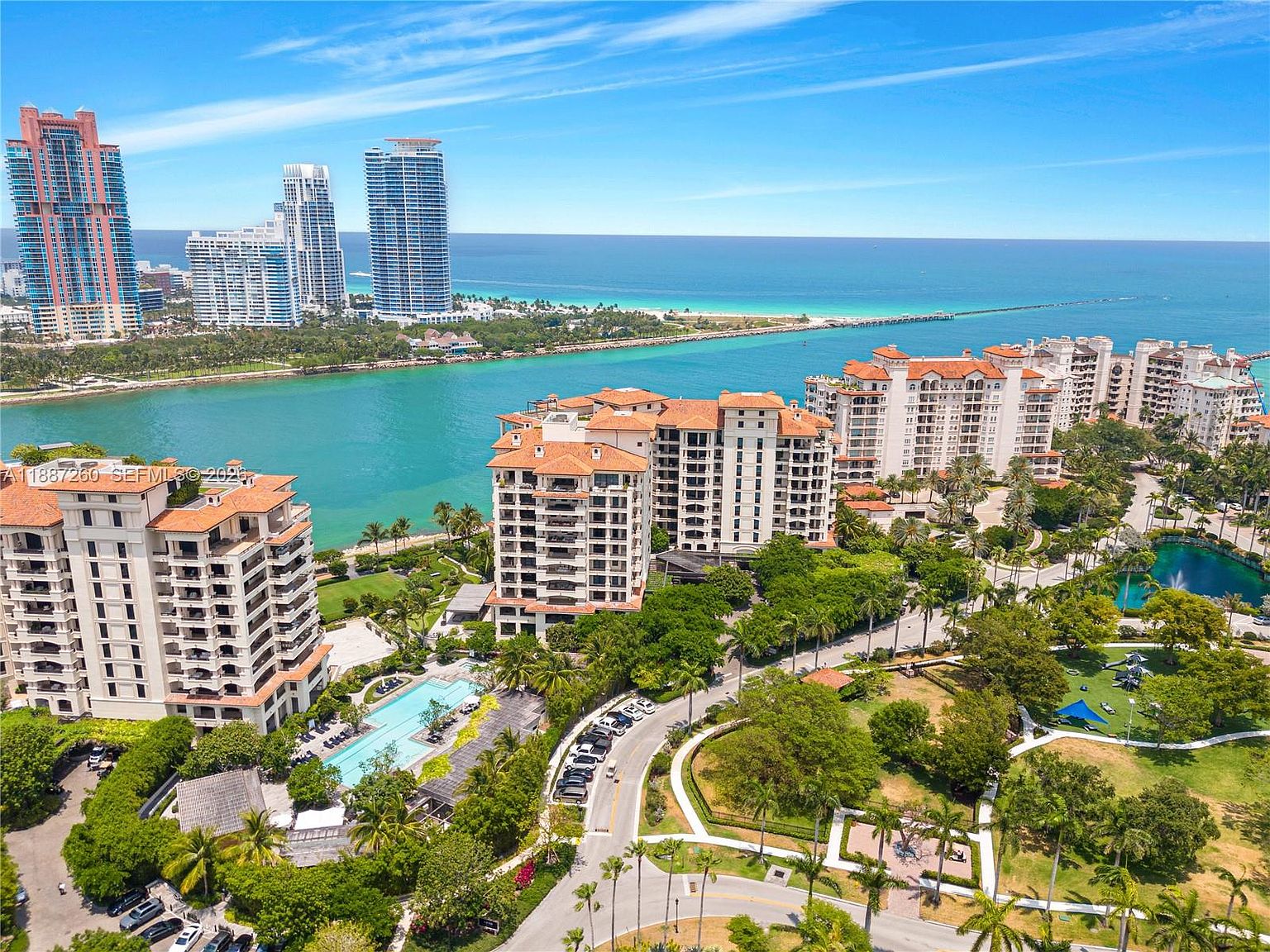 This high-angle aerial view captures a luxurious waterfront residential complex featuring multiple mid-rise buildings with terracotta-tiled roofs. The property is surrounded by lush tropical landscaping, a swimming pool, and winding pathways, all set against the backdrop of a vibrant blue bay and distant city skyline. The perspective emphasizes the resort-style atmosphere and the prime coastal location of the community.