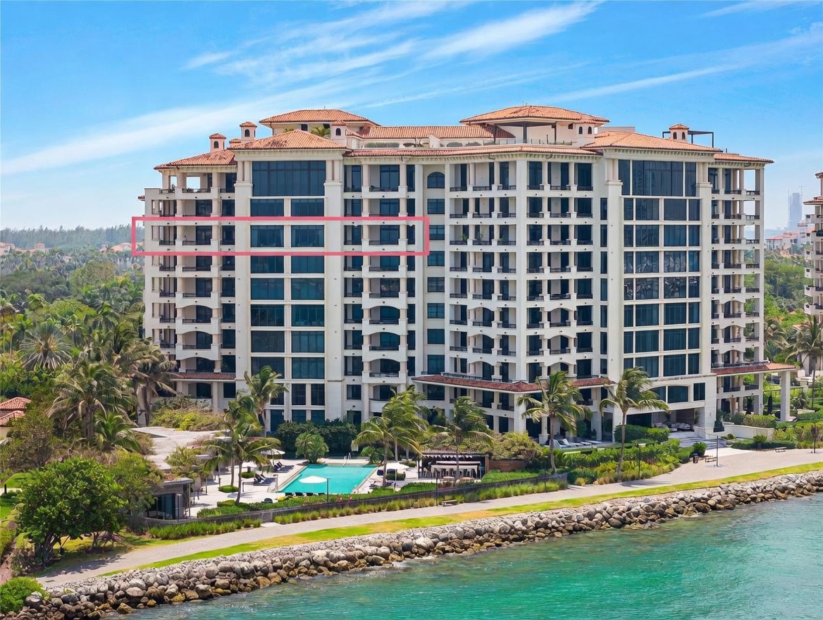 This aerial perspective captures a luxurious, multi-story waterfront residential building featuring a classic Mediterranean-inspired architectural style with a tiled roof. The property boasts extensive amenities, including a pristine swimming pool area surrounded by lush landscaping and palm trees, all situated directly along a scenic coastline. A pink rectangular overlay highlights a specific floor, emphasizing the building's expansive glass-fronted balconies and prime oceanfront location.