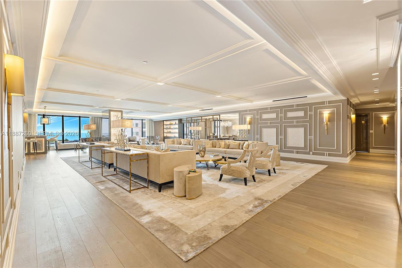 This expansive, high-end living room features a sophisticated neutral color palette with plush cream-colored sofas and elegant accent chairs arranged on a large, textured area rug. The room is defined by a coffered ceiling with recessed lighting, polished light-wood flooring, and decorative wall paneling that adds a sense of architectural depth. Large floor-to-ceiling windows provide an abundance of natural light and a view of the outdoors, creating an airy and luxurious atmosphere.