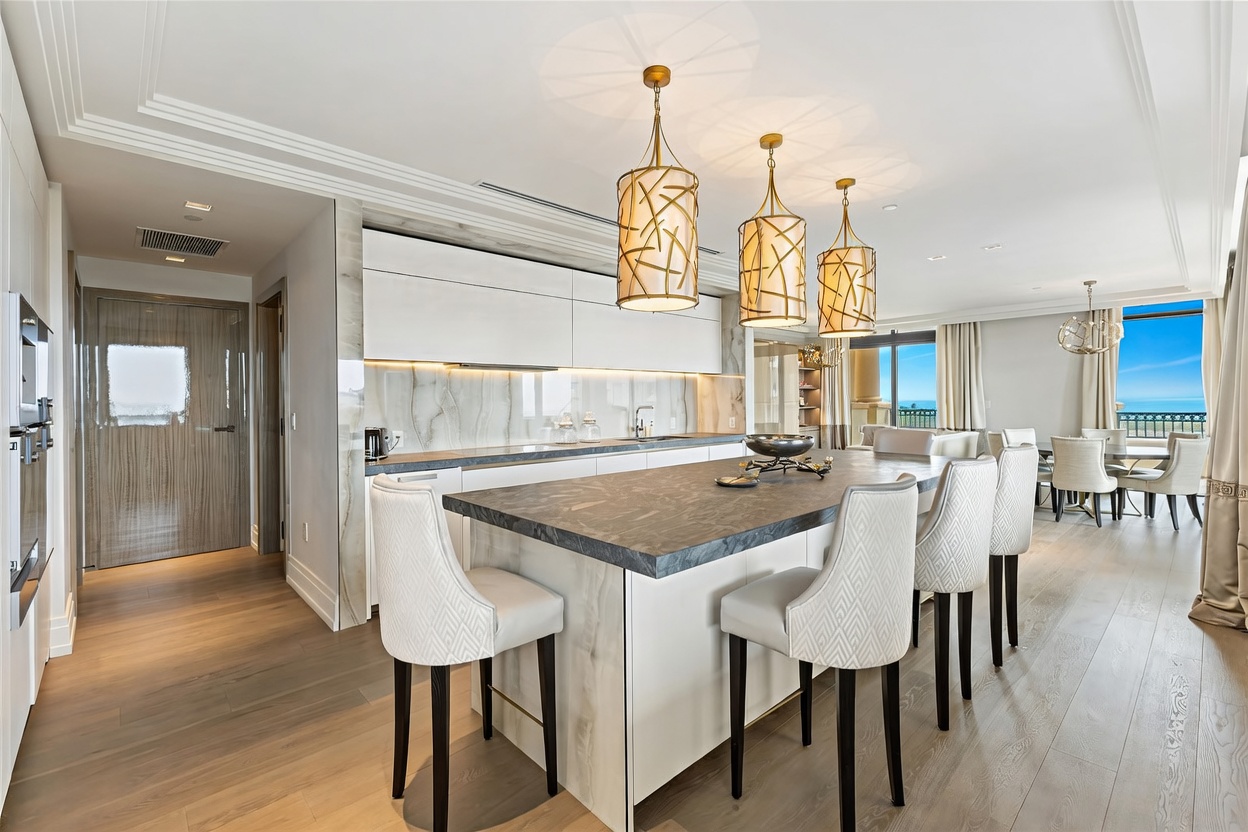 This sophisticated, modern kitchen features a large central island with a dark stone countertop and four elegant, upholstered bar stools. The space is highlighted by three ornate gold pendant lights, sleek white cabinetry, and a stunning marble-patterned backsplash. The open-concept design flows seamlessly into a dining area with a view of the ocean, creating a bright and luxurious atmosphere.