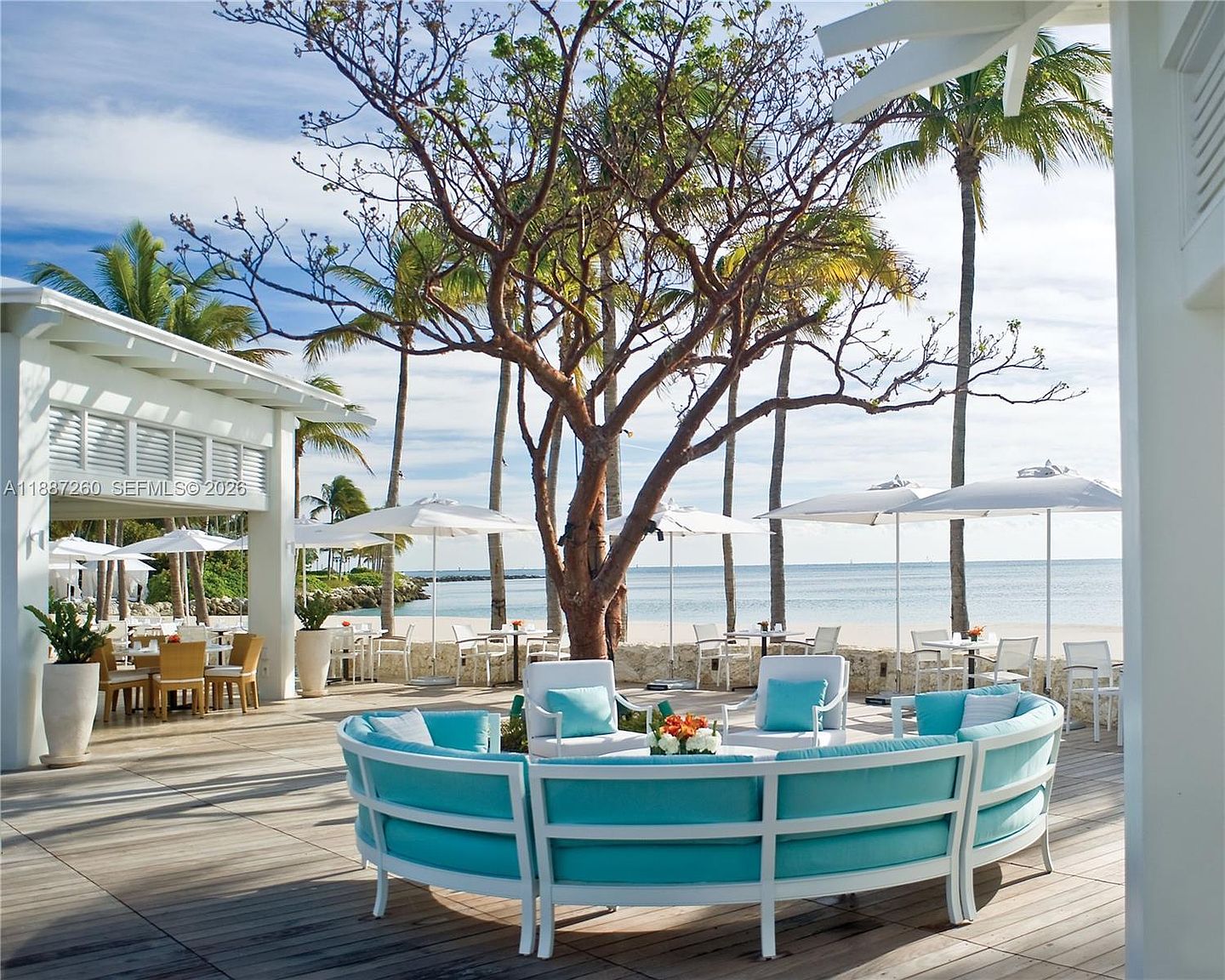 This expansive, sun-drenched outdoor deck features a luxurious, curved lounge seating area with vibrant turquoise cushions, perfect for relaxing by the ocean. The space is framed by mature trees and white umbrellas, offering a seamless transition from the covered patio area to the sandy beach and turquoise waters beyond. The overall atmosphere is one of high-end coastal elegance and serene tropical relaxation.