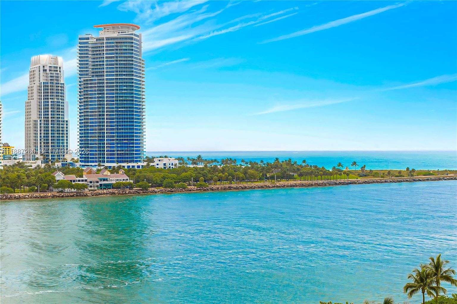 This wide-angle aerial view captures a stunning coastal landscape featuring two prominent, modern high-rise residential towers overlooking a vibrant turquoise bay. The foreground is dominated by the sparkling water, while the mid-ground showcases a lush green park area with palm trees and a walking path leading toward the ocean horizon. The scene conveys a luxurious, serene, and tropical lifestyle, emphasizing the prime waterfront location of the property.