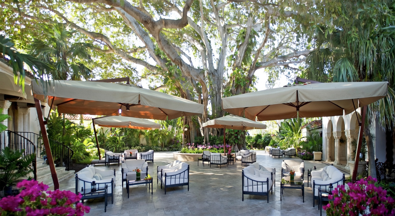 This expansive outdoor patio area features multiple large, beige cantilever umbrellas providing shade over several comfortable seating arrangements with white cushioned furniture. The space is surrounded by lush tropical landscaping, vibrant pink bougainvillea, and a massive, mature tree that serves as a stunning natural canopy. The stone-paved terrace offers a luxurious, resort-like atmosphere perfect for entertaining in a serene garden setting.