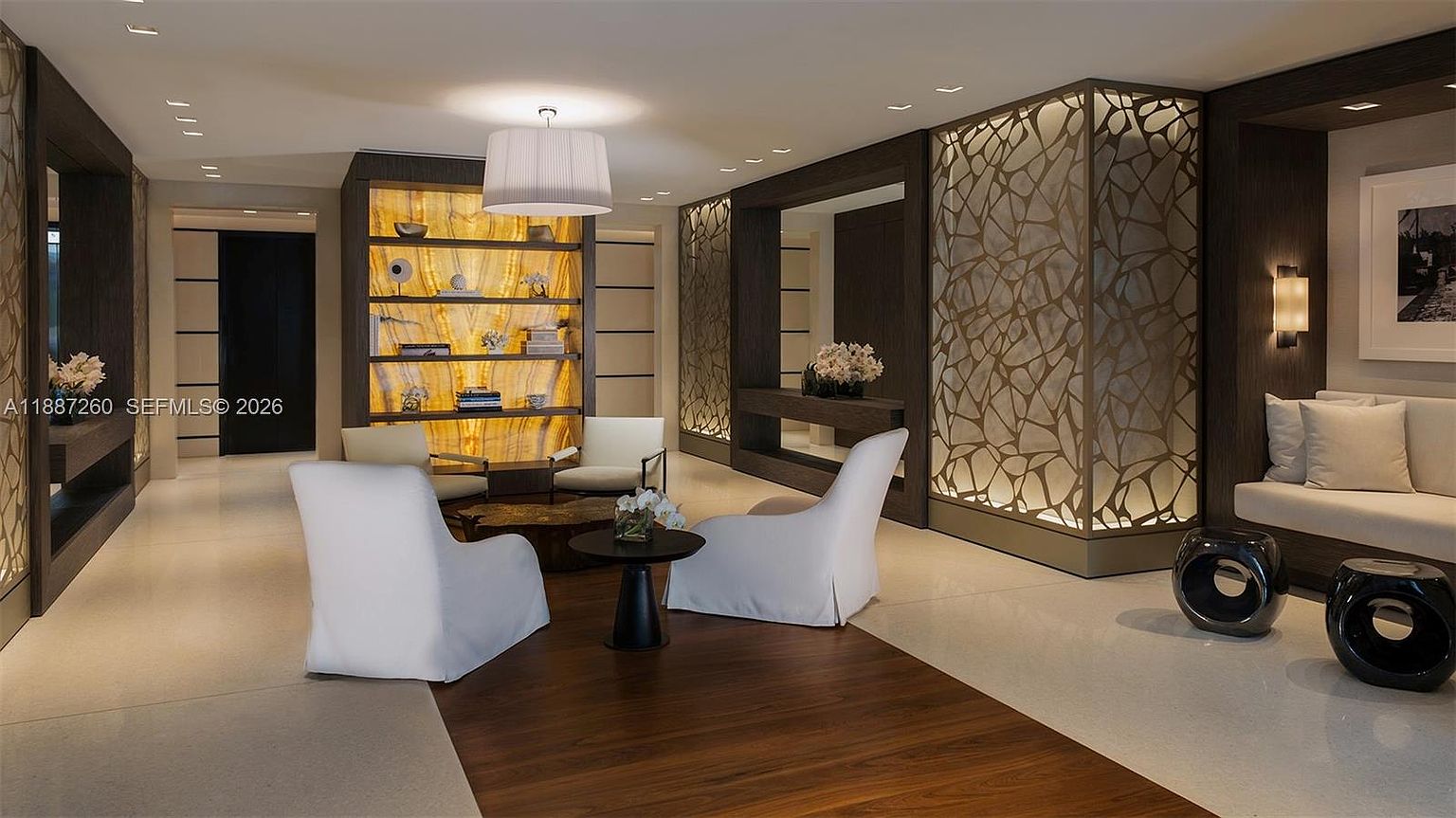 This sophisticated and modern living area features a warm, backlit onyx shelving unit as a focal point, complemented by elegant white armchairs and a dark wood floor inlay. The space is defined by high-end finishes, including intricate metallic room dividers and recessed lighting, creating a luxurious and tranquil atmosphere. The perspective is a wide-angle shot that captures the open-concept layout, emphasizing the refined textures and contemporary design elements.