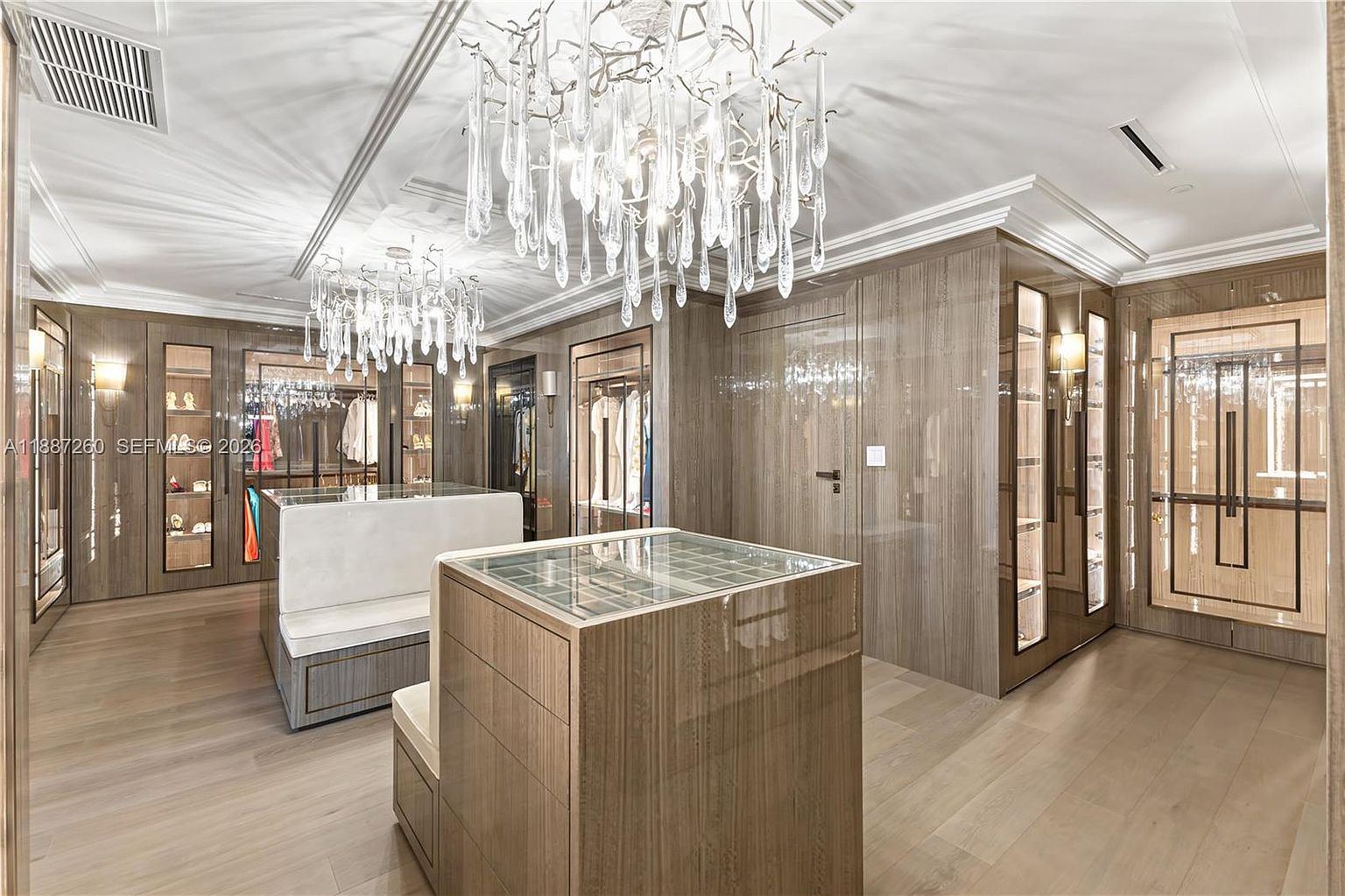 This luxurious, high-end walk-in closet features custom glossy wood cabinetry, integrated glass-front display cases, and two elegant crystal chandeliers that serve as the focal point. A central island with a glass top provides a sophisticated display area, while light-toned hardwood flooring adds warmth to the polished, modern aesthetic. The perspective is a wide-angle shot from the entrance, showcasing the expansive storage capacity and refined design of this boutique-style dressing room.