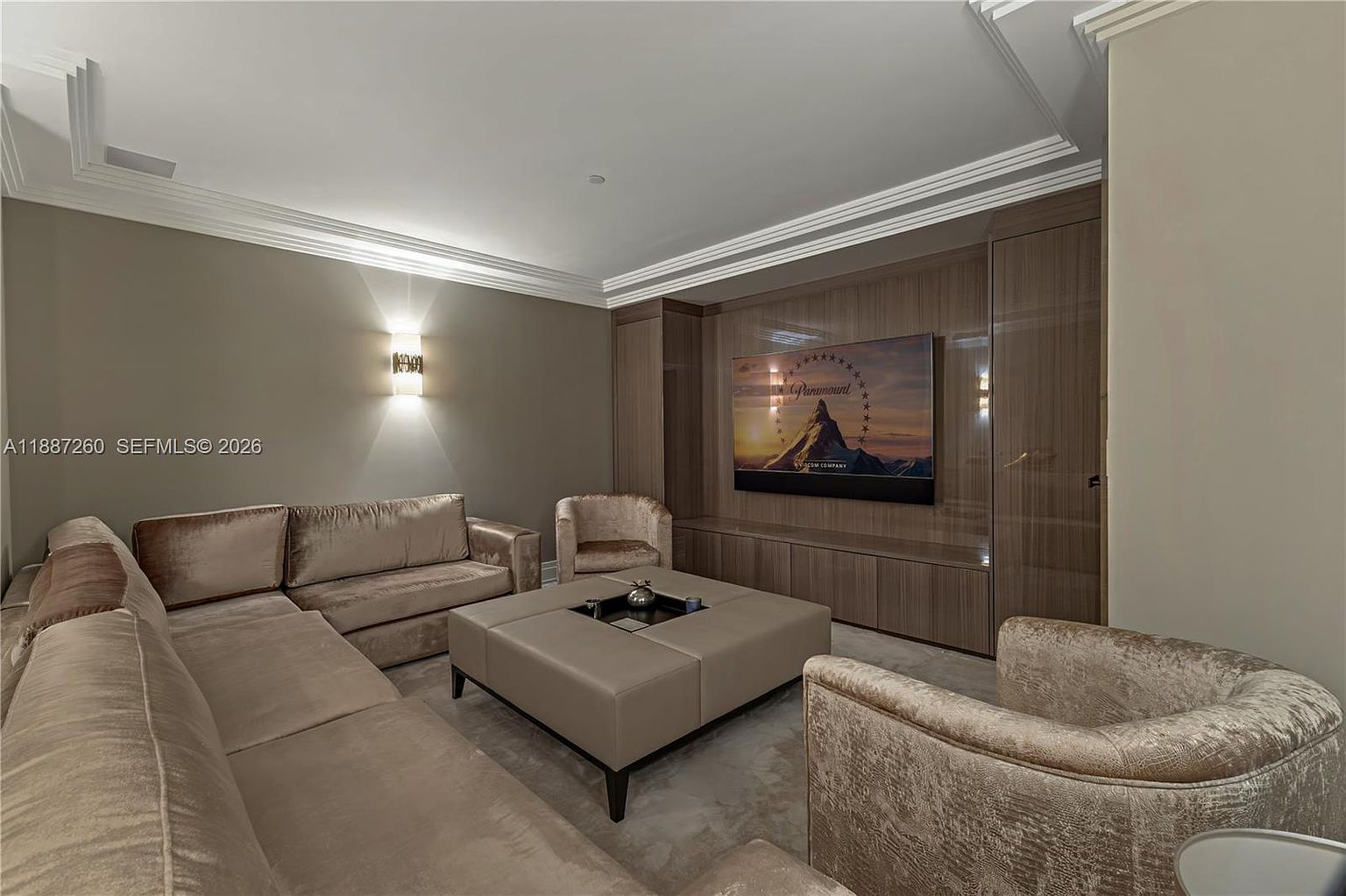 This sophisticated living room features a large, plush sectional sofa and two matching velvet armchairs arranged around a central leather ottoman. The focal point is a wall-mounted television set against a sleek, wood-paneled media wall with integrated cabinetry. The room is bathed in warm, ambient lighting from wall sconces, creating an inviting and luxurious atmosphere.