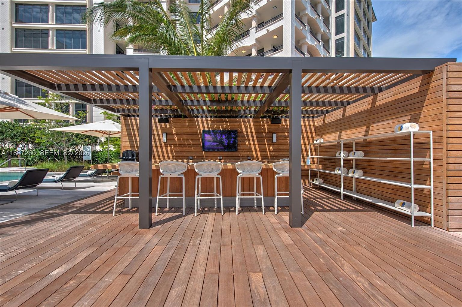 This image showcases a sophisticated outdoor lounge area featuring a wooden pergola structure with a built-in bar and television. The space is furnished with modern white bar stools and includes a shelving unit stocked with rolled towels, suggesting a luxury pool or spa amenity. The perspective is a wide, eye-level shot that emphasizes the warm wood tones and the inviting, resort-style atmosphere.