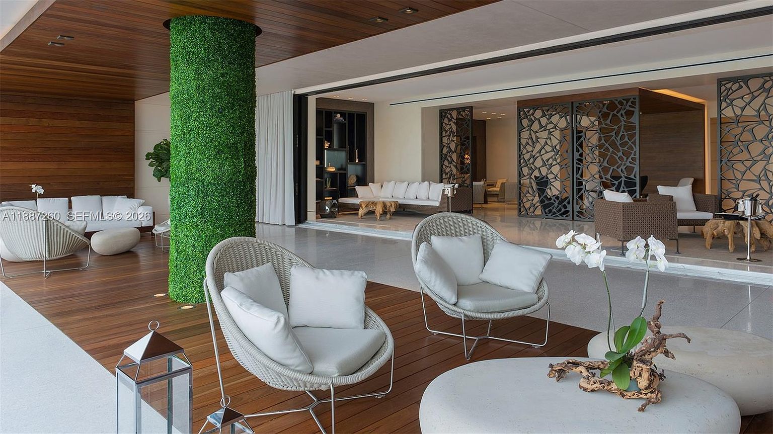 This luxurious, expansive covered patio features a seamless transition between indoor and outdoor living spaces, highlighted by a striking, moss-covered cylindrical pillar. The area is furnished with modern, light-toned wicker seating and sleek, minimalist furniture, set against a backdrop of warm wood ceilings and sophisticated decorative metal screens. The open-concept design creates an airy, resort-like atmosphere perfect for high-end entertaining.