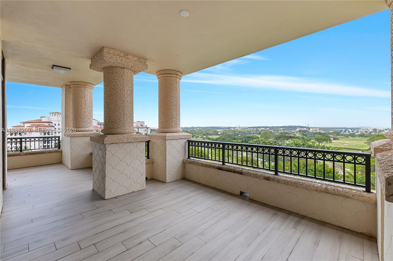 This spacious, covered balcony features light-toned wood-look tile flooring and elegant stone-textured columns, creating a sophisticated outdoor living space. The area is enclosed by a decorative black metal railing that offers an expansive, unobstructed view of a lush green golf course and distant residential buildings. The bright, open-air design provides a serene atmosphere, perfect for enjoying the panoramic landscape.