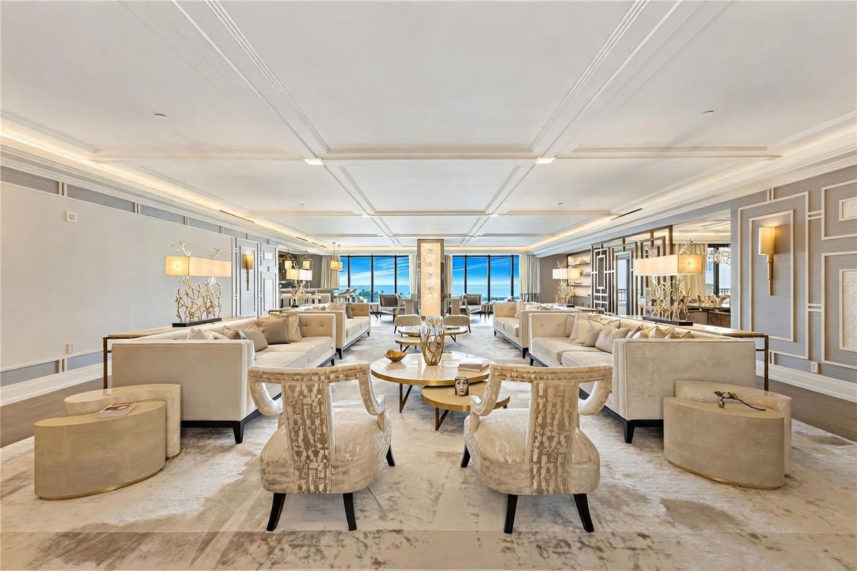 This expansive, symmetrical living room exudes luxury with its coffered ceiling, neutral color palette, and high-end contemporary furnishings. Two large, plush sofas face each other across a central circular coffee table, framed by elegant accent chairs and floor-to-ceiling windows that offer a stunning panoramic ocean view. The space is meticulously designed with sophisticated lighting fixtures and a refined, airy atmosphere perfect for grand entertaining.