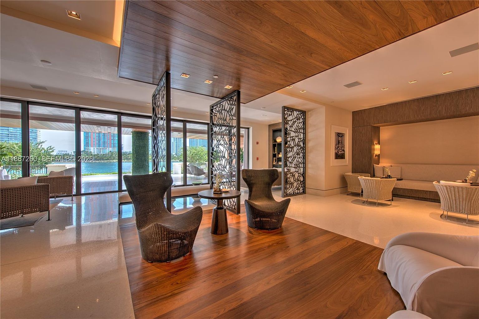 This sophisticated lounge area, likely part of a luxury building's amenity space, features a seamless blend of indoor and outdoor living with floor-to-ceiling glass walls overlooking a waterfront. The room is elegantly furnished with modern wicker armchairs, a central pedestal table, and a plush sofa area, all set against a rich wood-paneled ceiling and decorative metal room dividers. The overall aesthetic is upscale and tranquil, offering a perfect space for relaxation or social gatherings.