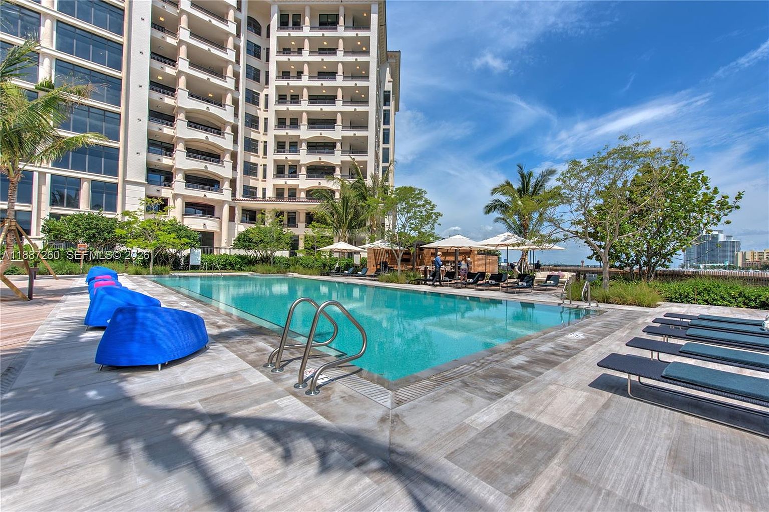 This image captures a serene, resort-style swimming pool area set against a tall, multi-story residential building. The scene features vibrant blue lounge chairs, sleek dark sun loungers, and white umbrellas, all arranged on a modern tiled deck. The perspective is at eye level, emphasizing the inviting turquoise water and the tranquil atmosphere of this luxury community amenity.