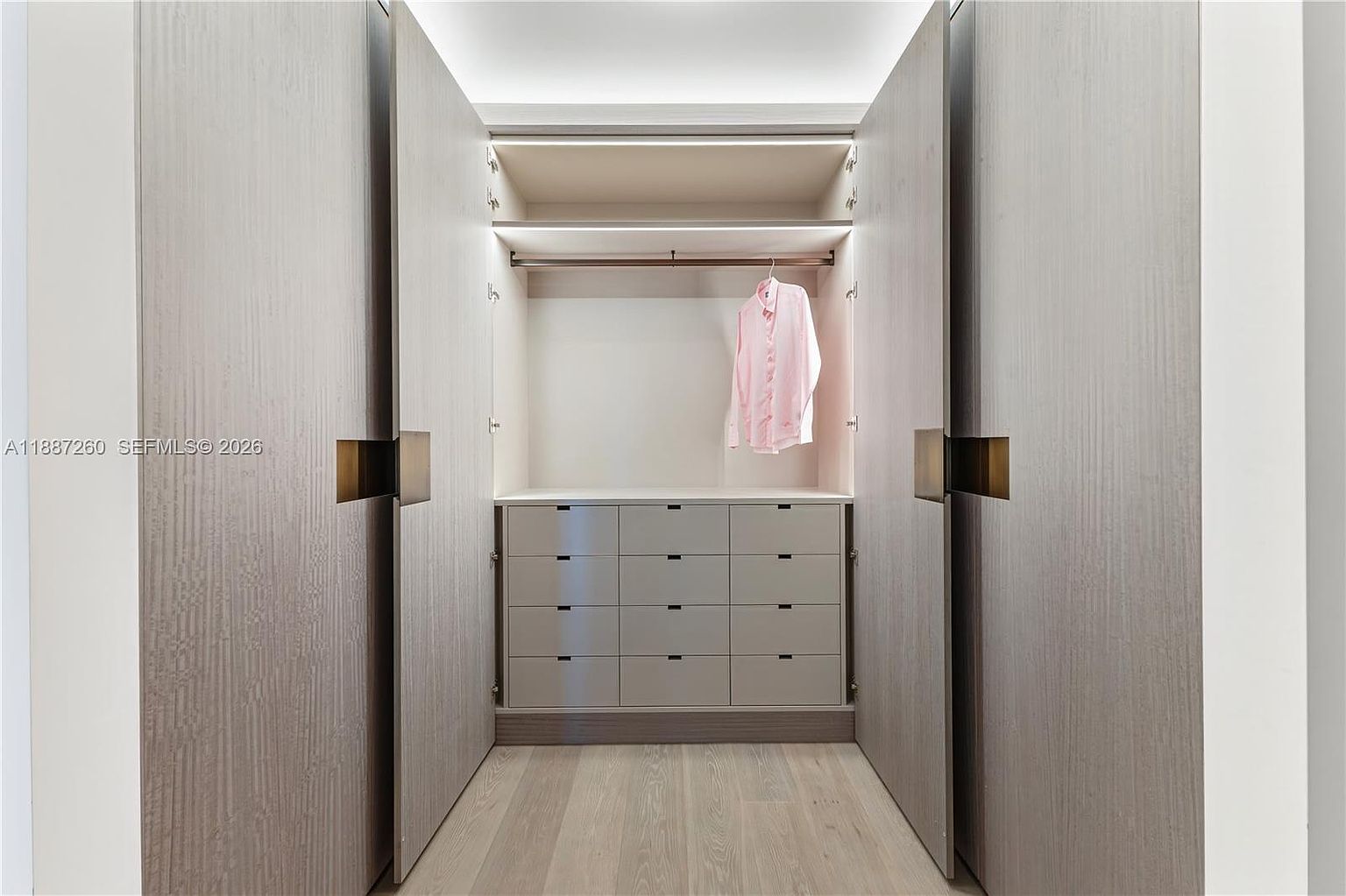 This modern, minimalist walk-in closet features sleek, light-wood paneled doors that open to reveal a well-organized interior. The space includes a hanging rod with a single garment, integrated LED lighting, and a built-in dresser with nine glossy drawers. The light-toned wood flooring and clean lines create a sophisticated and airy atmosphere.