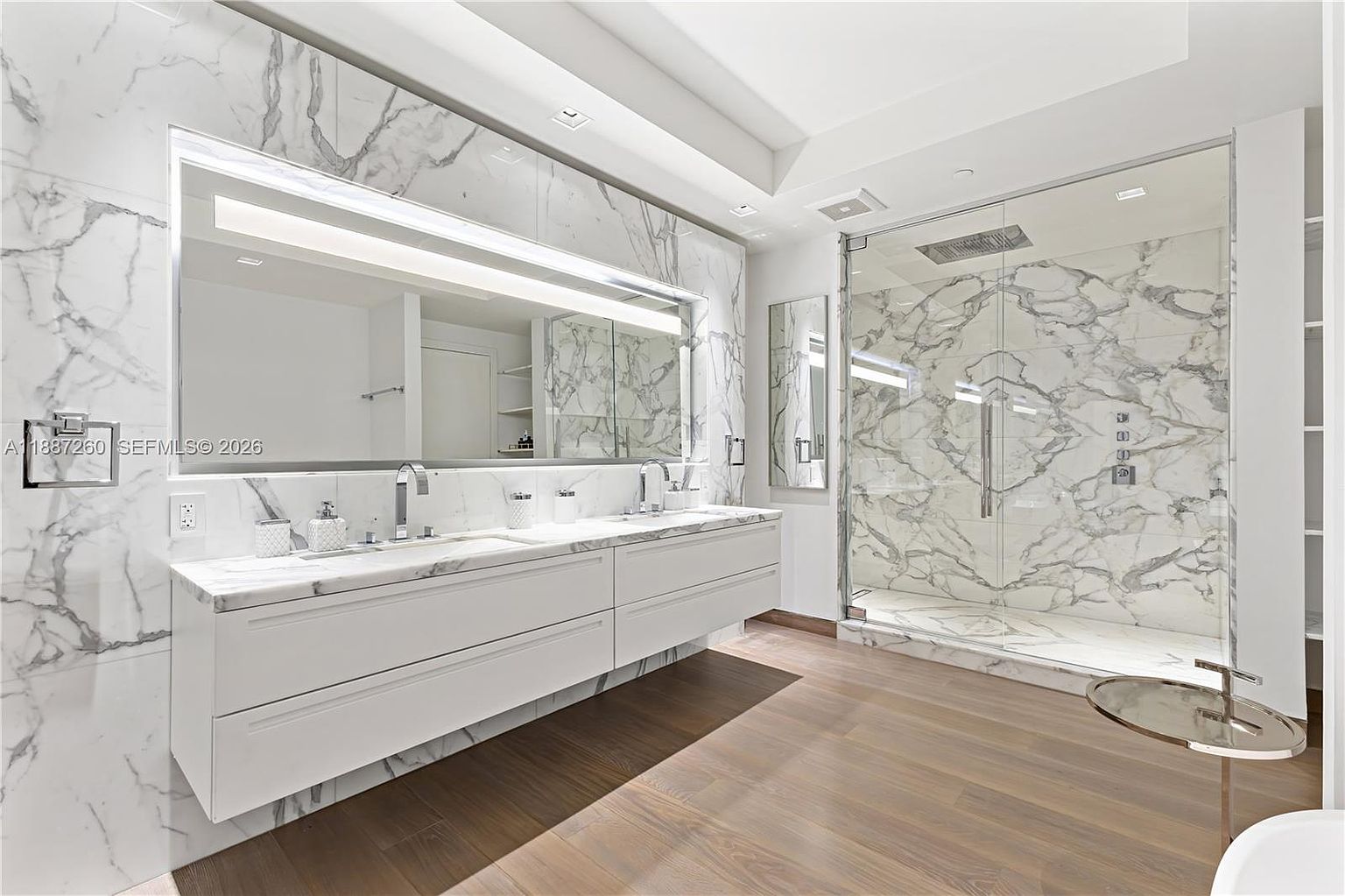 This luxurious primary bathroom features a sleek, floating double vanity with white cabinetry and marble countertops, set against a dramatic floor-to-ceiling marble wall with an integrated mirror. To the right, a spacious walk-in glass shower showcases matching marble tile work and high-end fixtures. The room is grounded by warm-toned wood flooring, creating a sophisticated and contemporary spa-like atmosphere.