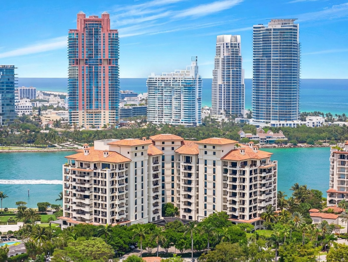 This high-angle aerial shot captures a prominent, multi-story residential building with a terracotta-tiled roof, situated amidst lush tropical landscaping and overlooking a serene bay. In the background, a striking skyline of modern glass skyscrapers rises against a clear blue sky, emphasizing the property's prime coastal location. The composition highlights the contrast between the Mediterranean-inspired architecture of the foreground building and the sleek, contemporary urban development of the surrounding area.