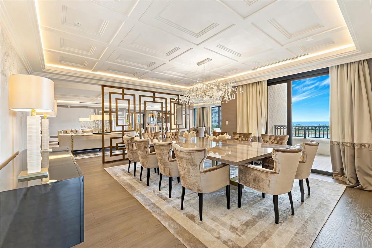 This elegant dining room features a large, polished table surrounded by plush, velvet-upholstered chairs, set beneath a stunning crystal chandelier. A decorative geometric room divider separates the dining area from the living space, while floor-to-ceiling windows offer a bright, airy atmosphere with a view of the ocean. The room is finished with sophisticated coffered ceilings and a neutral-toned area rug, creating a luxurious and refined aesthetic.