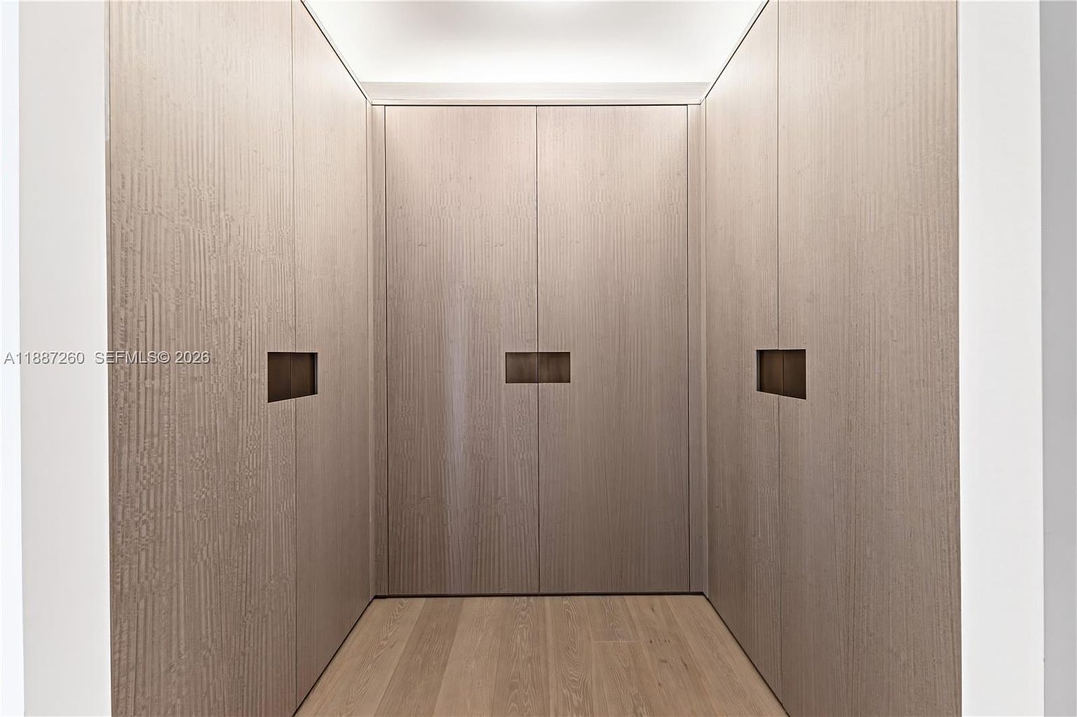 This image showcases a sleek, modern walk-in closet featuring floor-to-ceiling wood-paneled cabinetry with a minimalist design. The space is illuminated by recessed cove lighting at the ceiling, creating a warm and sophisticated atmosphere. The perspective is a straight-on, symmetrical shot that emphasizes the clean lines and high-end finishes of the storage area.