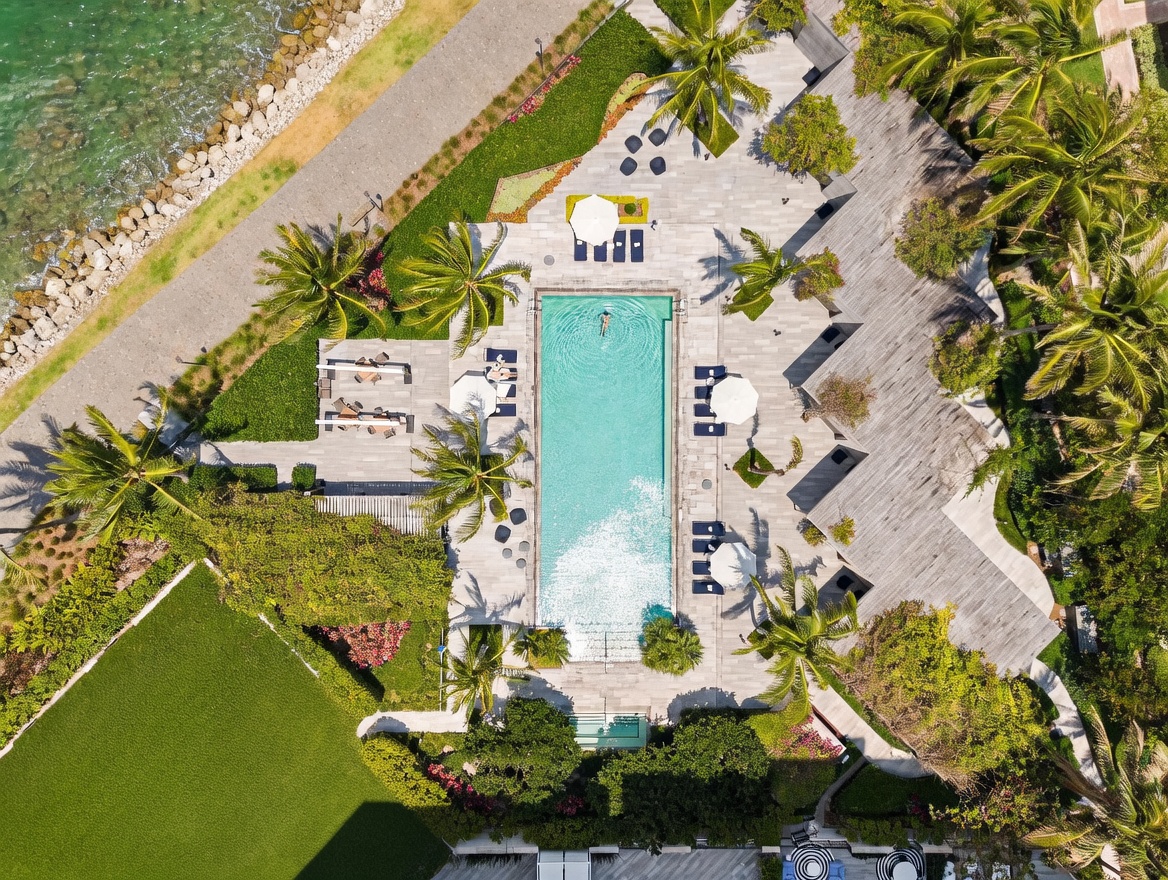 This stunning aerial view captures a luxurious resort-style swimming pool area surrounded by lush tropical landscaping and palm trees. The scene features a long, rectangular pool with a swimmer, multiple lounge chairs with umbrellas, and a wooden deck, all situated near a rocky shoreline. The perspective provides a comprehensive look at the outdoor amenities, highlighting the serene and upscale atmosphere of the property.