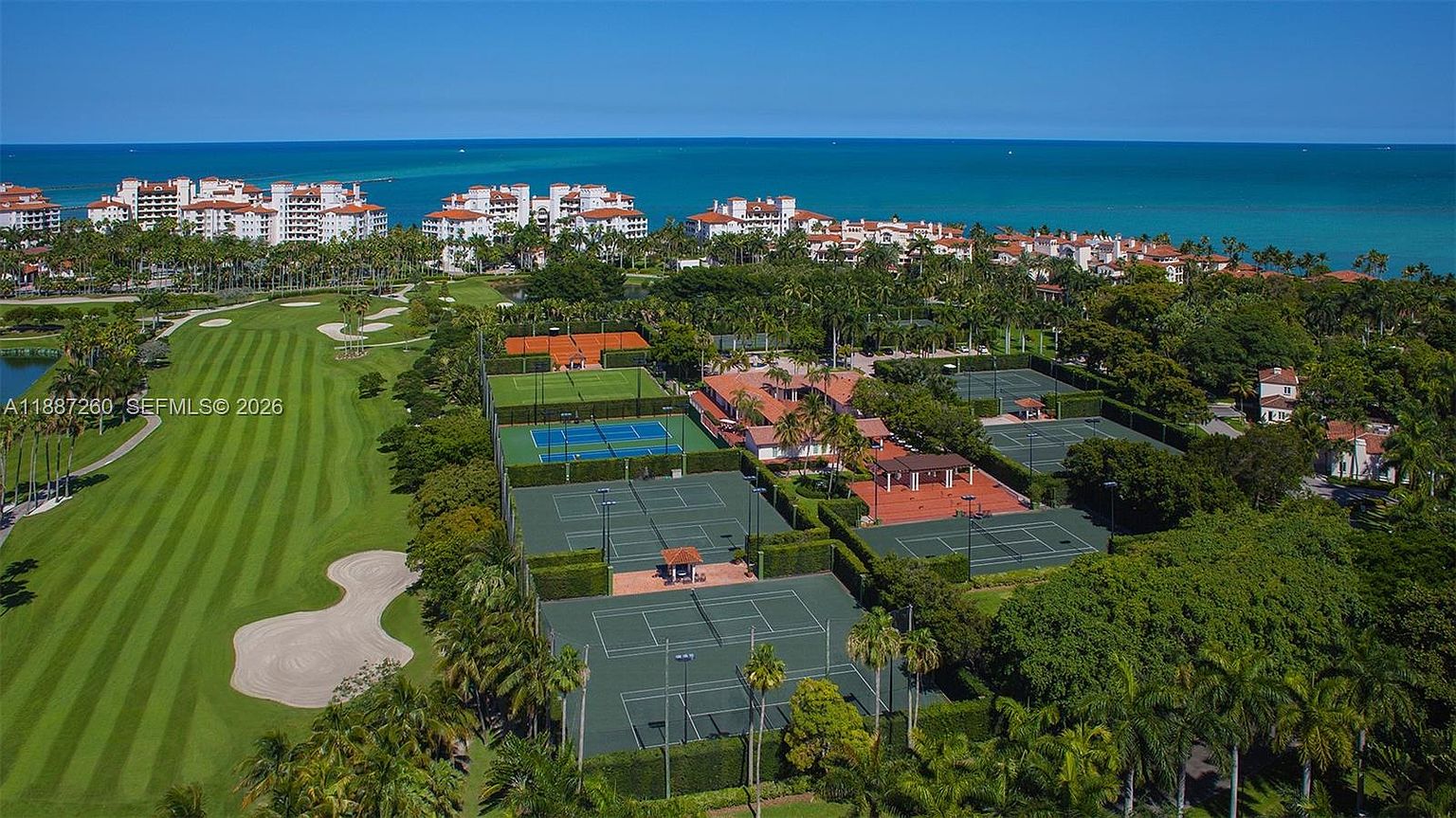 This high-angle aerial view captures a sprawling luxury residential community featuring a pristine golf course, multiple tennis courts, and lush tropical landscaping. In the background, large Mediterranean-style residential buildings overlook the turquoise ocean, creating a resort-like atmosphere. The perspective emphasizes the expansive scale and premium recreational amenities available within this exclusive coastal neighborhood.