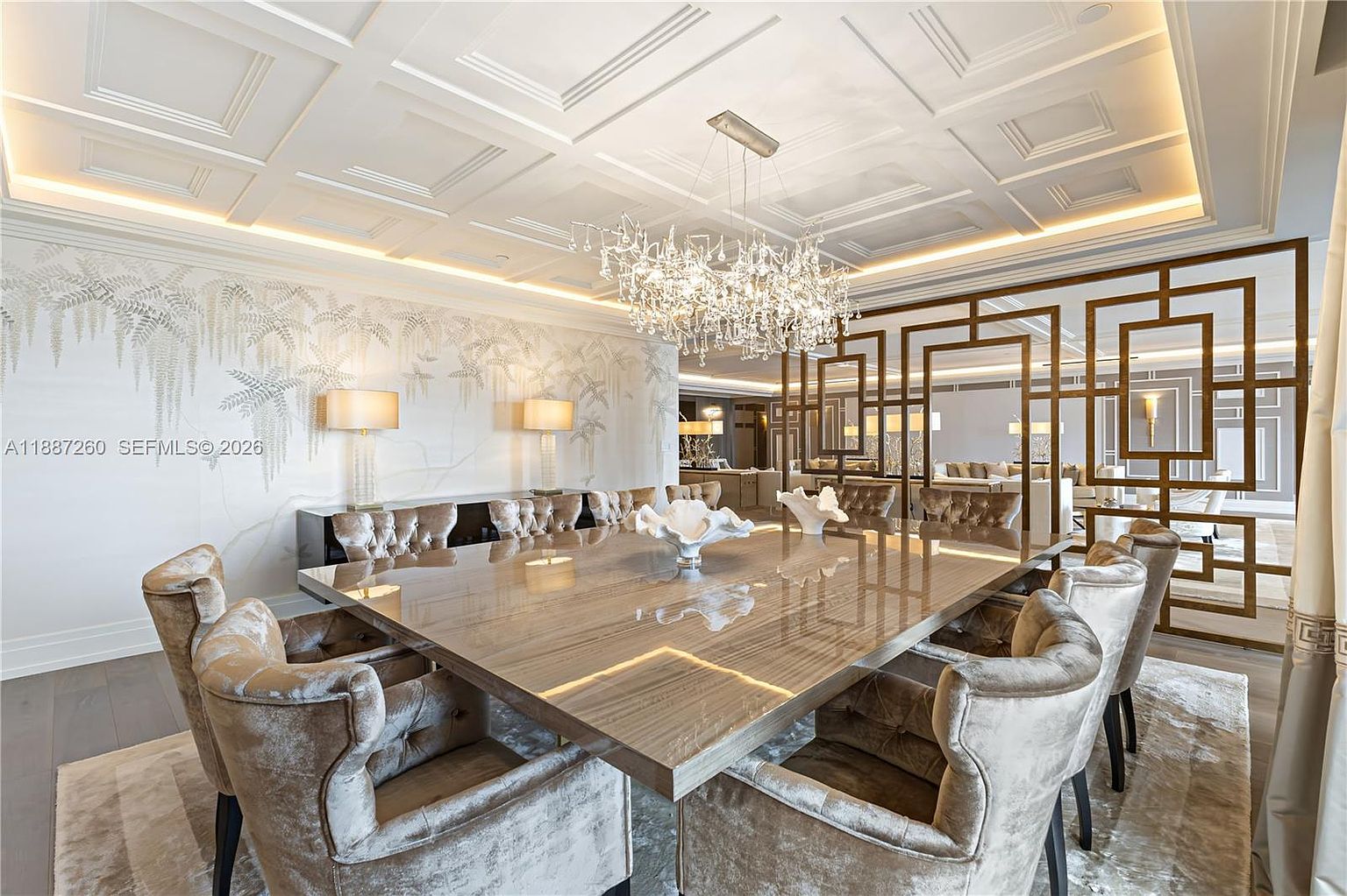 This elegant dining room features a large, polished wood table surrounded by plush, velvet-upholstered chairs, centered under a dramatic, multi-tiered crystal chandelier. The room is defined by a sophisticated coffered ceiling, a delicate fern-patterned wallpaper, and a geometric gold-toned room divider that adds a touch of modern luxury. The perspective is eye-level, capturing the opulent atmosphere and the seamless flow into the adjacent living space.