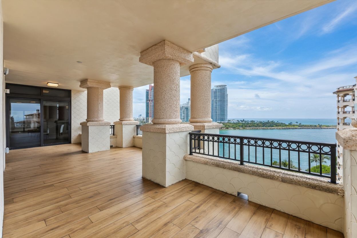 This expansive, covered balcony features elegant stone columns and light-toned wood-look tile flooring, creating a sophisticated outdoor living space. The area is framed by a decorative black metal railing and offers breathtaking, unobstructed panoramic views of the ocean and distant city skyline. The perspective is wide and inviting, emphasizing the seamless transition between indoor luxury and the serene coastal environment.