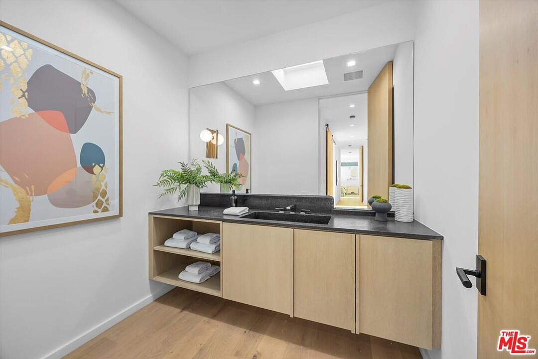 This modern powder room features a sleek, light wood vanity with an integrated stone sink and a dark stone countertop. A large, frameless mirror reflects the adjacent hallway, while a piece of abstract art adds a pop of color to the crisp white walls. The space is illuminated by a skylight and contemporary wall-mounted lighting, creating a bright and minimalist aesthetic.