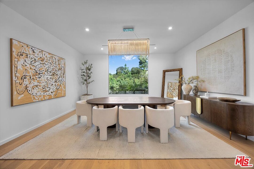 This elegant dining room features a large, dark wood oval table surrounded by six cream-colored upholstered chairs, all set atop a textured area rug. The space is anchored by a long, dark wood sideboard adorned with decorative vases and a large, framed abstract artwork on the wall, while a striking linear chandelier hangs above the table. A large window provides a view of lush greenery, and a tall floor mirror leans against the wall, enhancing the bright and sophisticated atmosphere.
