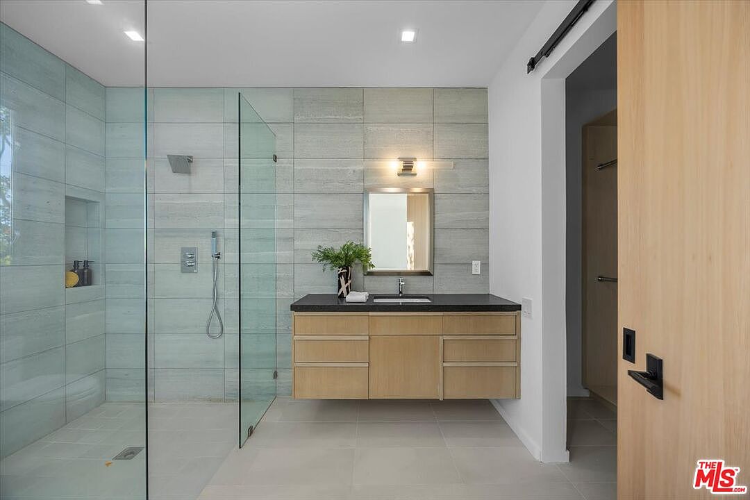 This modern bathroom features a floating light wood vanity with a dark countertop and a rectangular undermount sink. To the left, a spacious walk-in shower is enclosed by glass panels and finished with light-toned horizontal tile. The room is illuminated by a sleek wall-mounted light fixture above a rectangular mirror, creating a clean and contemporary aesthetic.