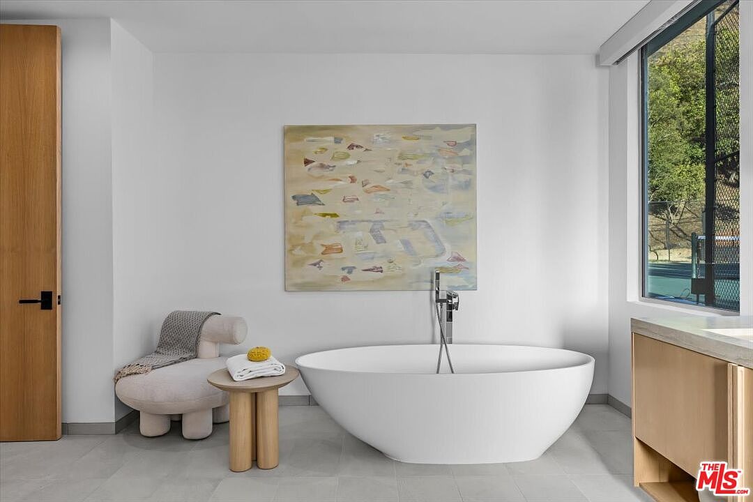 This modern bathroom features a sleek, white freestanding soaking tub positioned beneath a large piece of abstract wall art. To the left, a sculptural, light-colored armchair sits beside a small wooden side table, while a large window on the right provides natural light and a view of the outdoors. The space is characterized by a minimalist aesthetic with neutral tones and clean lines.