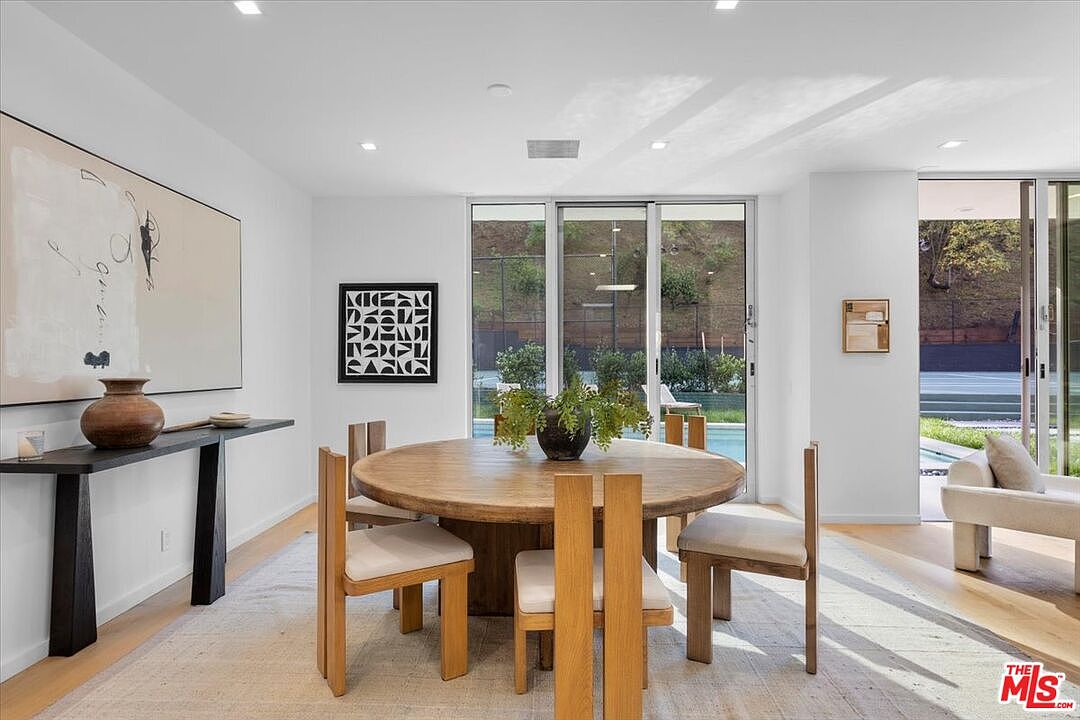 This bright and airy dining room features a round wooden table surrounded by minimalist light wood chairs, set atop a neutral area rug. A long, dark console table sits against the left wall beneath a large abstract painting, while a geometric black-and-white framed print hangs on the wall near the sliding glass doors. The space is illuminated by recessed lighting and offers a clear view through the large glass doors to the outdoor pool and landscaped hillside.