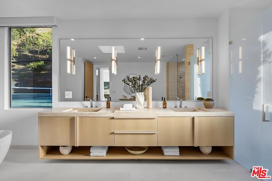 This modern primary bathroom features a long, light wood double vanity with integrated stone sinks and a large, wide mirror reflecting the room. The space is illuminated by four sleek, vertical wall-mounted light fixtures and includes an open lower shelf for storage. A large window to the left provides natural light and a view of an outdoor tennis court, creating a bright and airy atmosphere.