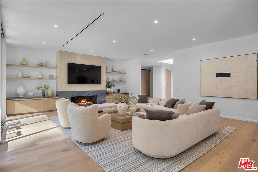 This spacious, contemporary living room features a neutral color palette with light wood flooring and a large area rug. A prominent stone-clad chimney breast serves as the focal point, housing a television above a fireplace, flanked by built-in light wood shelving. The room is furnished with a large, curved sectional sofa and two matching armchairs, creating an inviting and open-concept seating area.