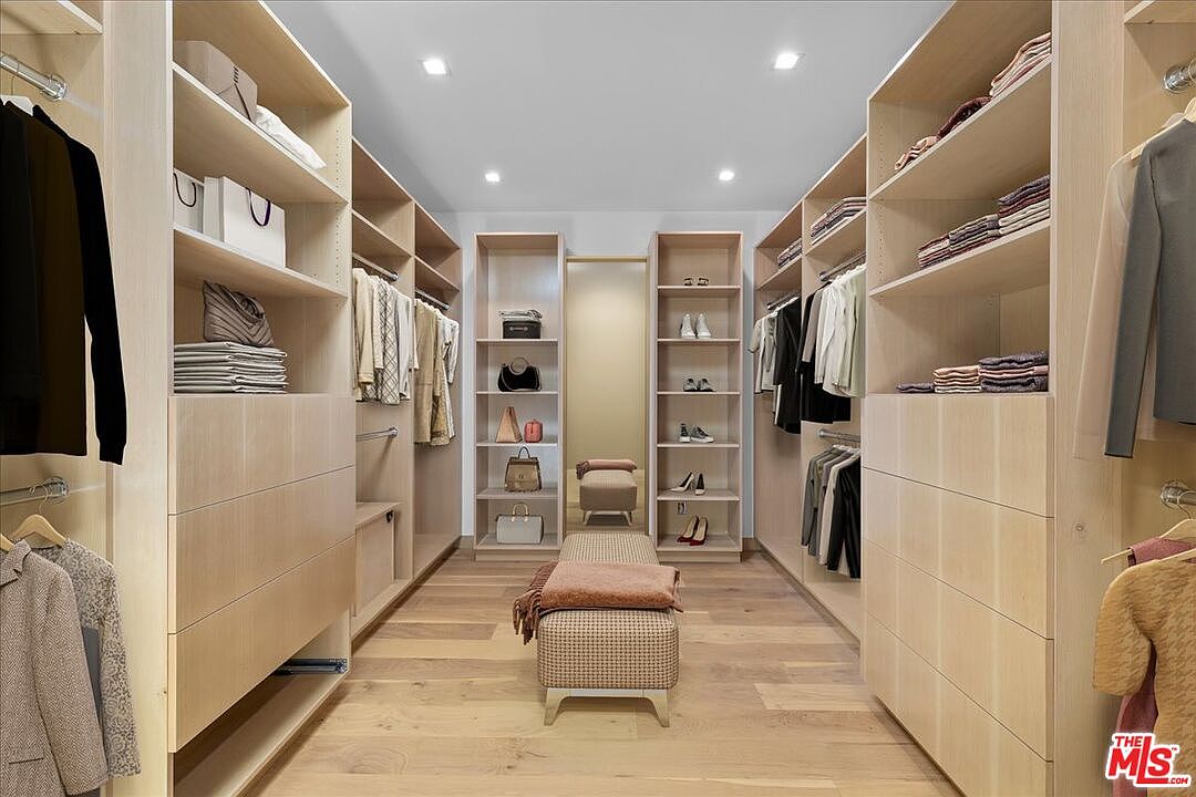 This spacious, well-organized walk-in closet features light wood cabinetry with extensive shelving, hanging rods, and built-in drawers. A central upholstered bench sits on the light wood flooring, providing a functional focal point in the middle of the room. The symmetrical layout and recessed ceiling lighting create a clean, high-end aesthetic suitable for a luxury primary suite.