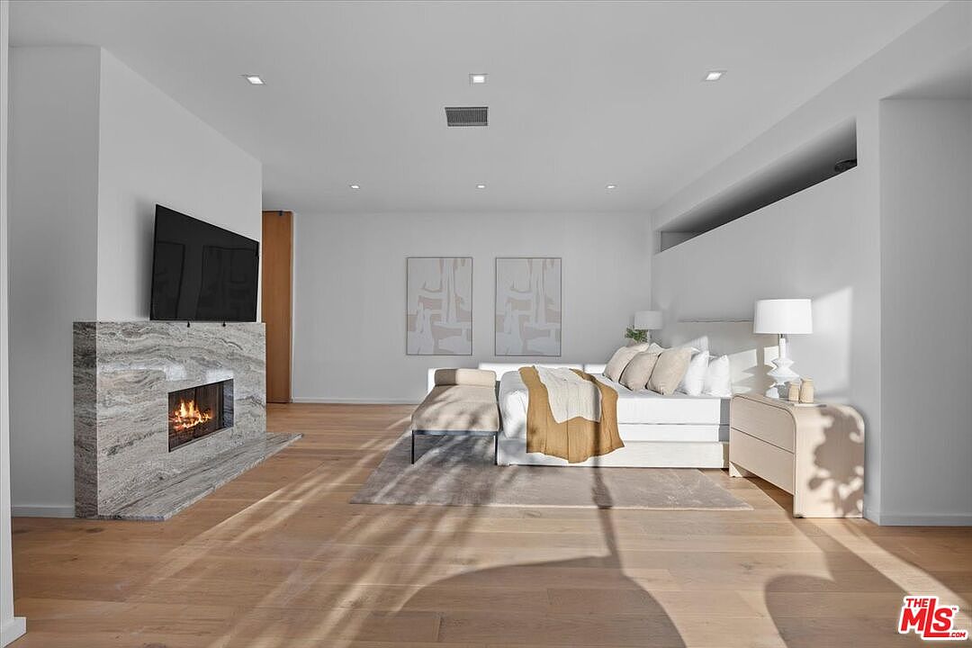 This spacious primary bedroom features light wood flooring and a neutral color palette, creating a serene and modern atmosphere. A prominent stone-clad chimney breast serves as a focal point, housing a fireplace and a wall-mounted television. The room is furnished with a plush bed, a bench at the foot, and a sleek bedside cabinet, all arranged to emphasize the open, airy layout.
