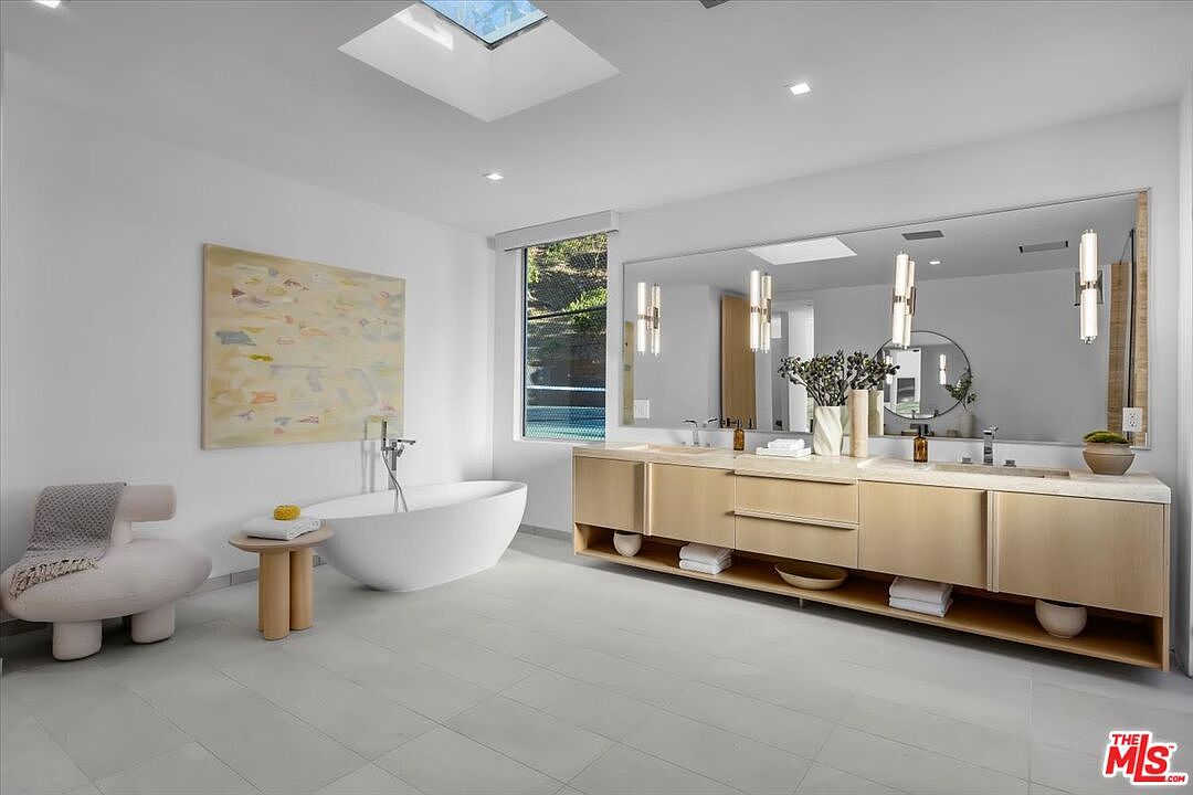This spacious, modern primary bathroom features a minimalist aesthetic with a freestanding soaking tub and a long, light wood vanity with dual sinks. A large, wall-to-wall mirror reflects the bright, airy space, which is illuminated by a skylight and contemporary pendant lighting. The room is finished with neutral-toned tile flooring and a soft, textured accent chair, creating a serene and luxurious atmosphere.