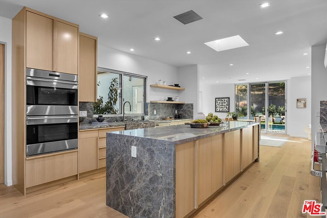 This modern kitchen features light wood cabinetry paired with a large central island topped with a dark, patterned stone surface. The space is brightened by recessed lighting and a skylight, while large sliding glass doors at the rear provide a seamless view of the outdoor pool area. The open-concept design flows into a living space, creating an airy and sophisticated atmosphere.
