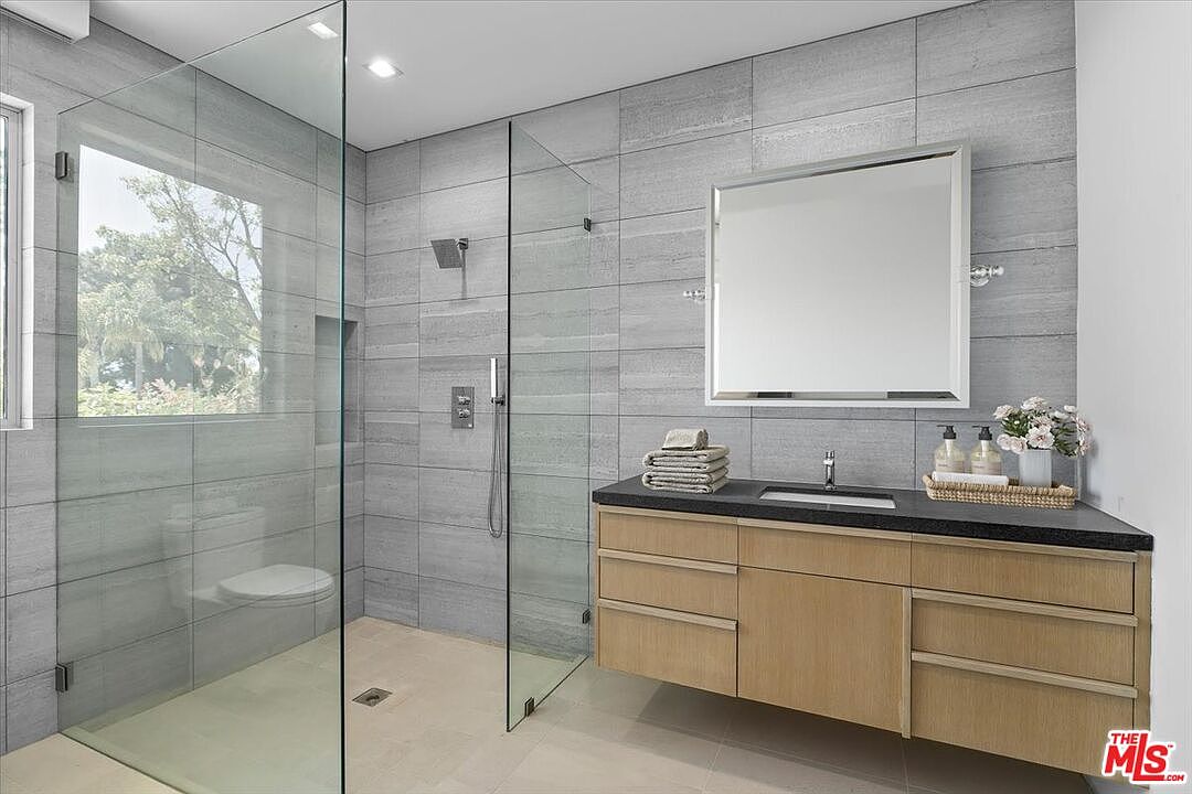 This modern bathroom features a walk-in shower enclosed by glass panels, showcasing light-toned, horizontal-patterned wall tiles. A floating vanity with light wood cabinetry and a dark stone countertop provides a clean, contemporary aesthetic, complemented by a rectangular mirror and minimalist fixtures. The space is bright and airy, with a window allowing natural light to filter into the shower area.