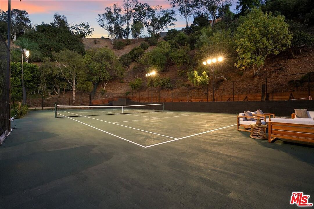 This image captures a private tennis court set against a lush, hillside backdrop during the golden hour. The court is illuminated by tall stadium-style floodlights, and a comfortable outdoor seating area with wooden furniture is positioned on the side for spectators. The scene conveys a sense of luxury and active outdoor living, framed by mature trees and a dark perimeter fence.
